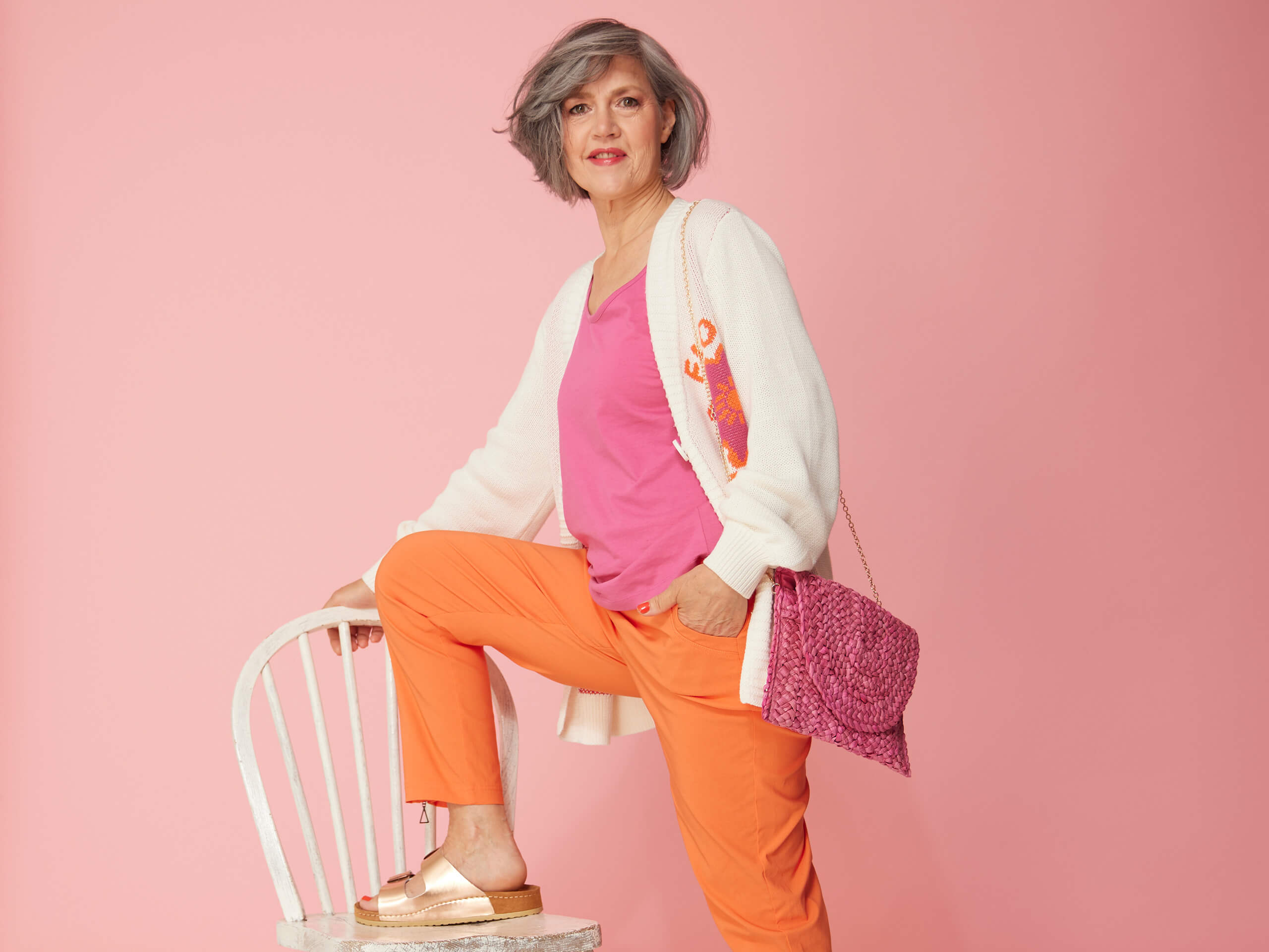 Ein Model trägt ein rosa Top, eine cremefarbene Strickjacke und eine orange Hose, steht mit einem Fuß auf einem weißen Stuhl vor rosa Hintergrund.