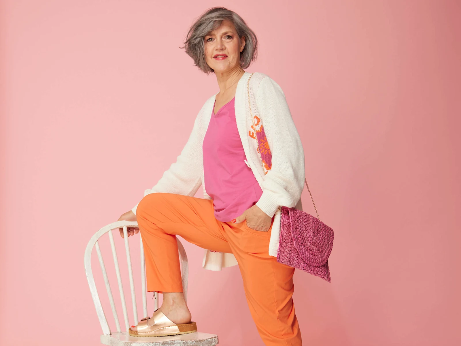 Ein Model trägt ein rosa Top, eine cremefarbene Strickjacke und eine orange Hose, steht mit einem Fuß auf einem weißen Stuhl vor rosa Hintergrund. Ein Model trägt ein rosa Top, eine cremefarbene Strickjacke und eine orange Hose, steht mit einem Fuß auf einem weißen Stuhl vor rosa Hintergrund.