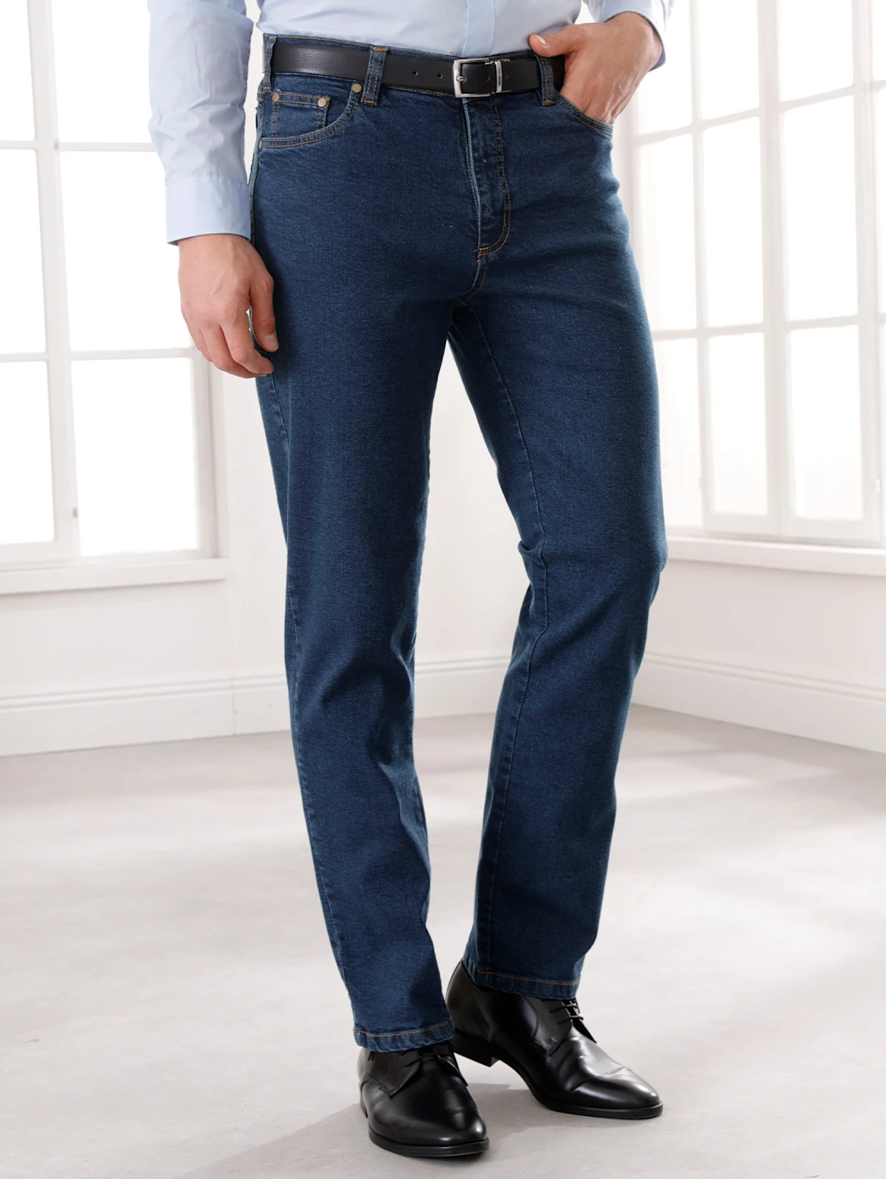 Mann trägt dunkelblaue Jeans, hellblaues Hemd, schwarzen Gürtel und schwarze Lederschuhe in einem hellen Raum mit großen Fenstern. Mann trägt dunkelblaue Jeans, hellblaues Hemd, schwarzen Gürtel und schwarze Lederschuhe in einem hellen Raum mit großen Fenstern.