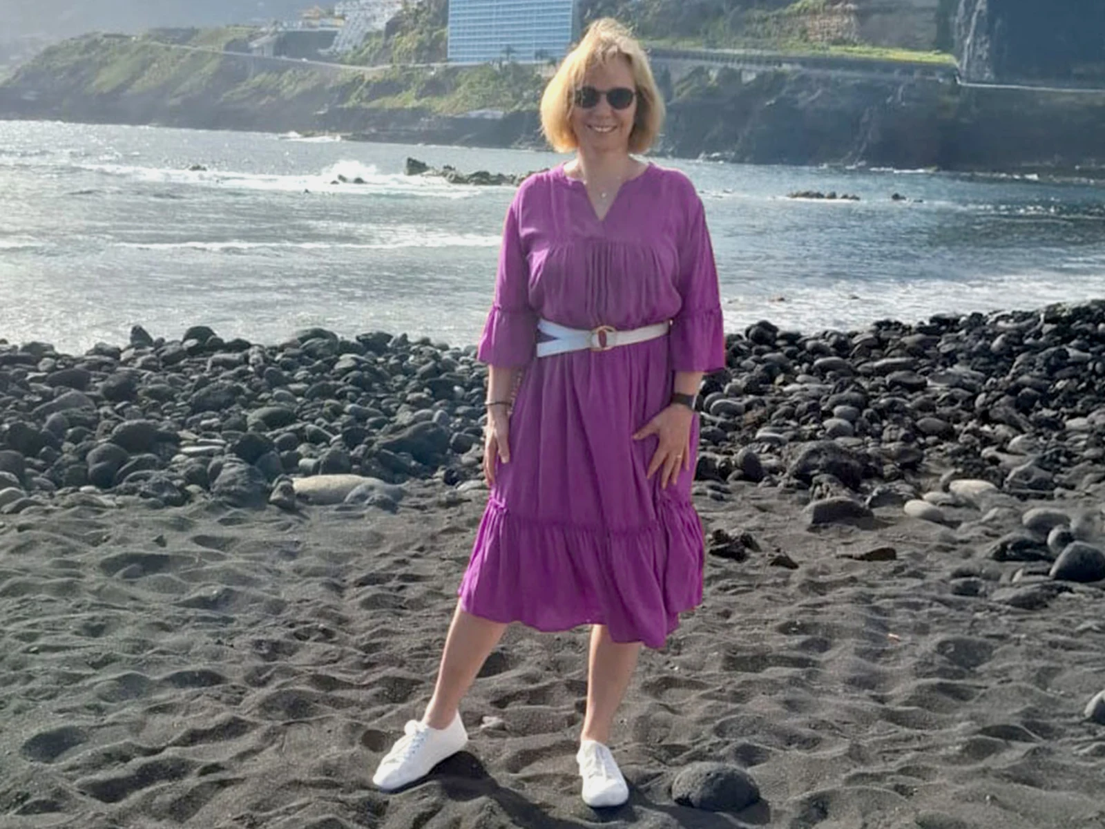 Eine Frau steht am Strand auf dunklem Sand, trägt ein lila Kleid mit Gürtel und weiße Schuhe. Im Hintergrund ist das Meer zu sehen. Eine Frau steht am Strand auf dunklem Sand, trägt ein lila Kleid mit Gürtel und weiße Schuhe. Im Hintergrund ist das Meer zu sehen.