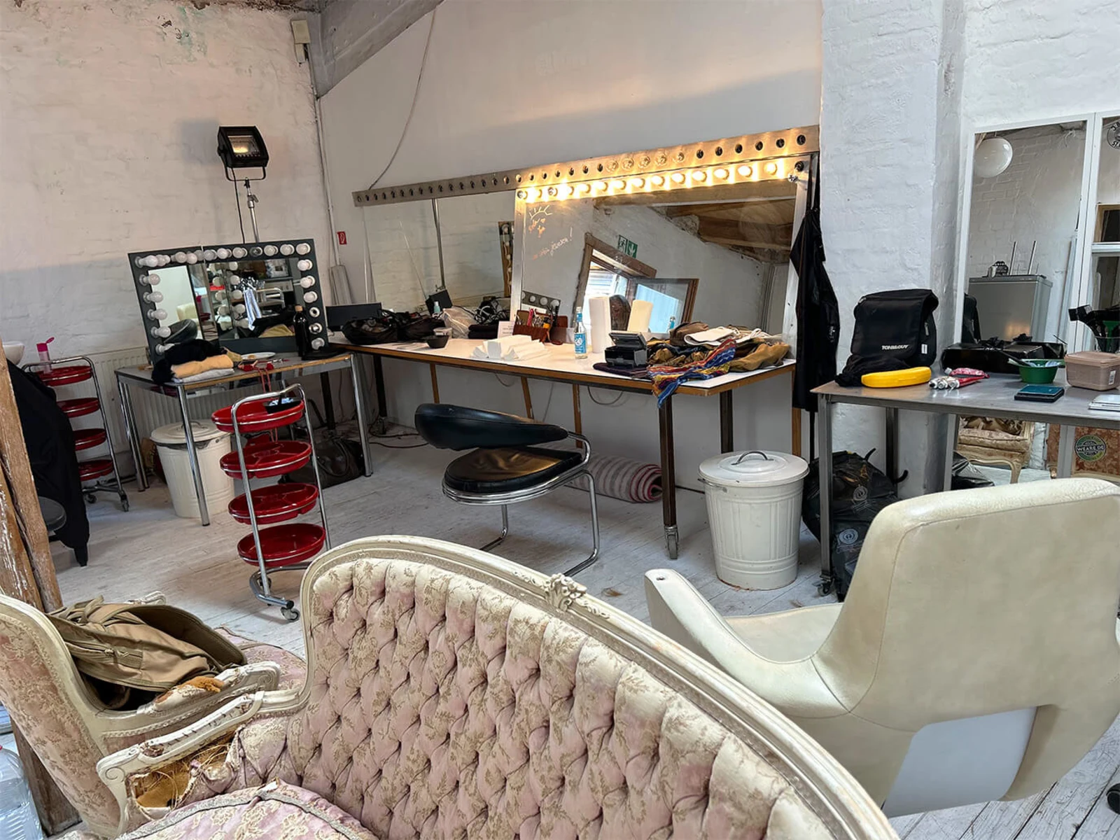 Ein Raum mit Vintage Sofa, Friseurstuhl, Spiegeln mit Lichtern und Tischen mit verschiedenen Gegenständen, helle Wände und Holzboden. Ein Raum mit Vintage Sofa, Friseurstuhl, Spiegeln mit Lichtern und Tischen mit verschiedenen Gegenständen, helle Wände und Holzboden.