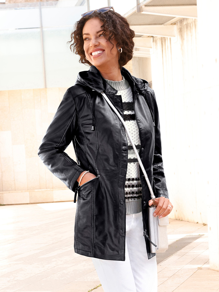 Ein Model trägt eine schwarze, lange Lederjacke mit Kapuze, darunter einen schwarz-weiß gemusterten Pullover und eine weiße Hose, hält eine weiße Tasche.