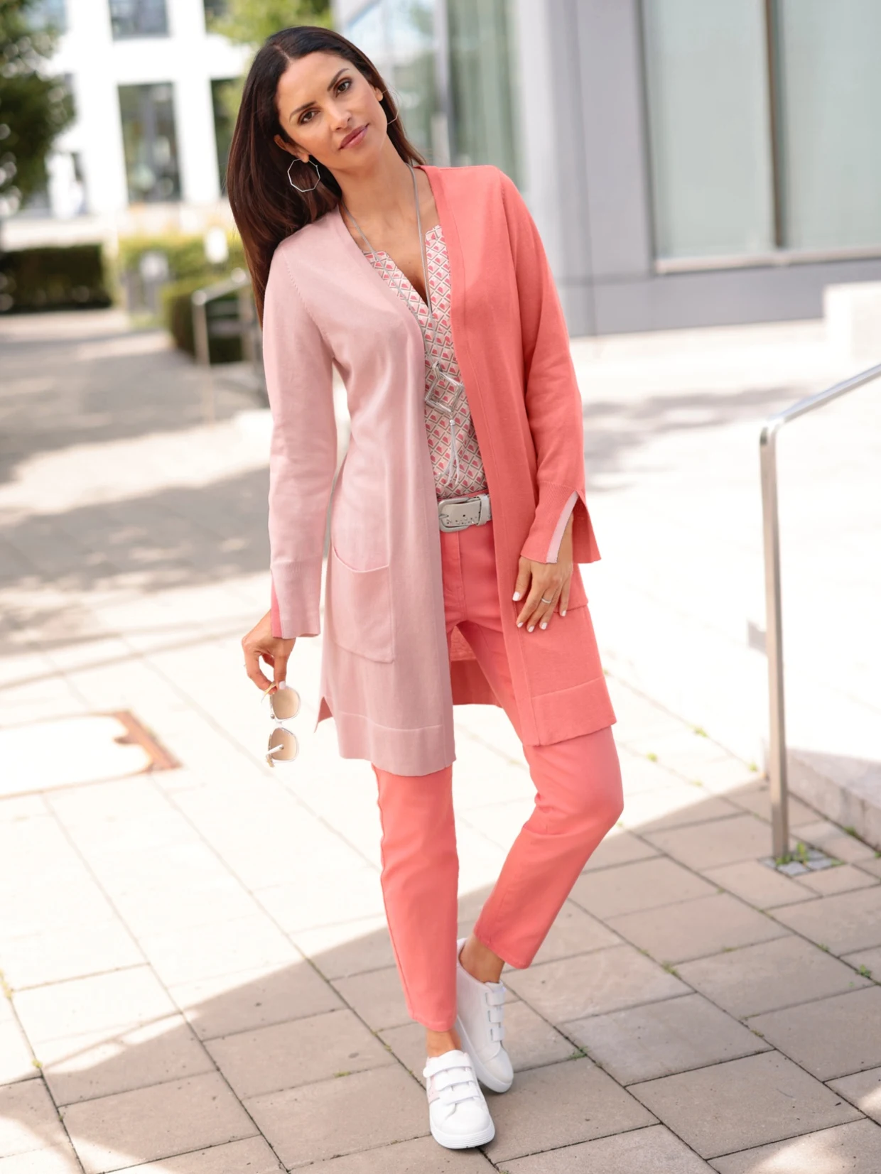 Frau trägt ein farblich abgestimmtes Outfit aus rosa Cardigan, gemusterter Bluse, korallfarbener Hose und weißen Sneakern, hält Sonnenbrille in der Hand, im Hintergrund moderne Architektur.