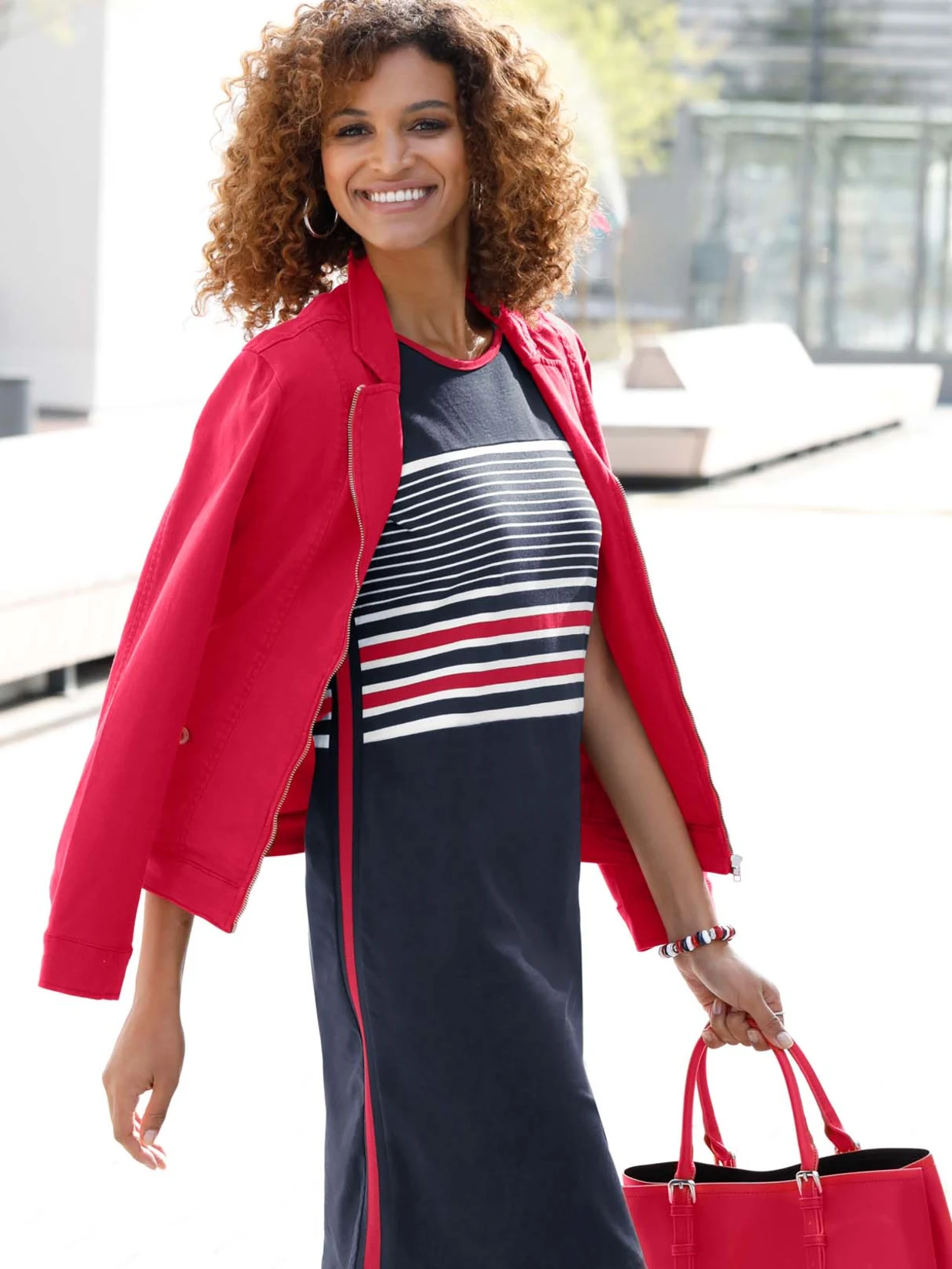 Frau trägt ein dunkelblaues Kleid mit weißen und roten Streifen, kombiniert mit einer roten Jacke und einer roten Handtasche.