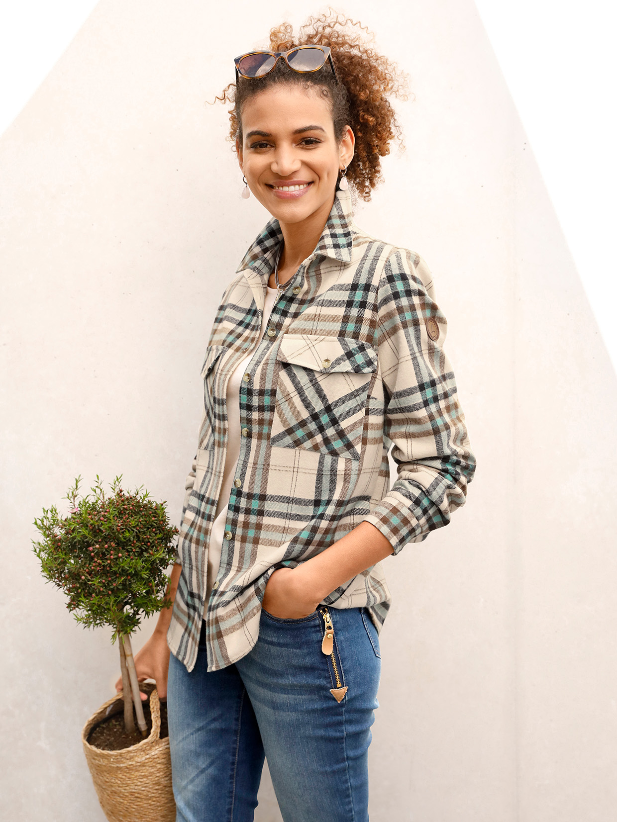 Karierte Damenbluse in Beige, Blau und Grün mit Brusttaschen, kombiniert mit blauen Jeans mit Reißverschlussdetail; im Hintergrund eine kleine Topfpflanze in einem Korb.