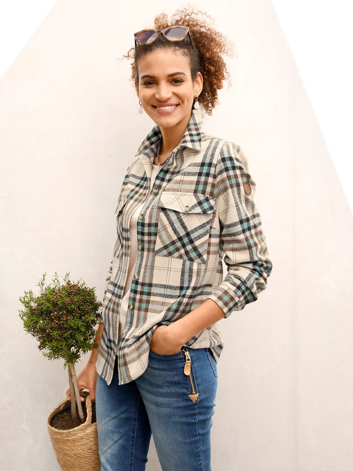 Karierte Damenbluse in Beige, Blau und Grün mit Brusttaschen, kombiniert mit blauen Jeans mit Reißverschlussdetail; im Hintergrund eine kleine Topfpflanze in einem Korb.