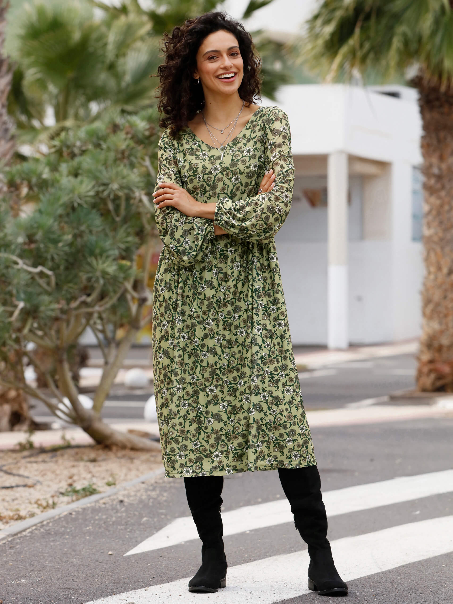 Ein Model trägt ein grünes, blumiges Kleid und schwarze Stiefel, steht in einer Umgebung mit Palmen.