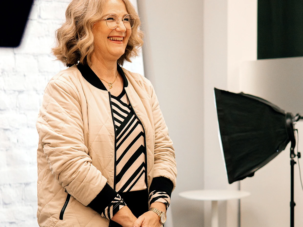 Eine lächelnde Frau mit Brille, beigem Stepp-Blouson und einem schwarz-beige gestreiften Oberteil steht vor einem weißen Hintergrund mit einem Softbox-Licht auf der rechten Seite. Eine lächelnde Frau mit Brille, beigem Stepp-Blouson und einem schwarz-beige gestreiften Oberteil steht vor einem weißen Hintergrund mit einem Softbox-Licht auf der rechten Seite.