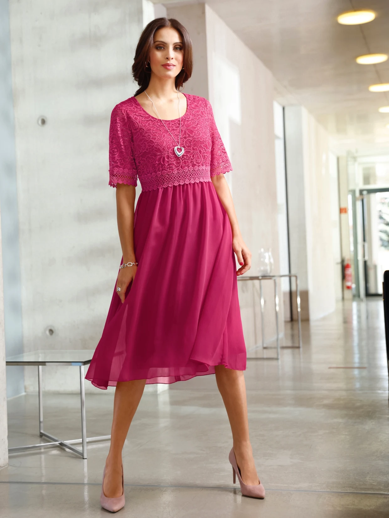 Frau trägt ein elegantes fuchsiafarbenes Kleid mit Spitzenoberteil und halbtransparentem Rock, kombiniert mit einer herzförmigen Halskette und nudefarbenen Pumps.