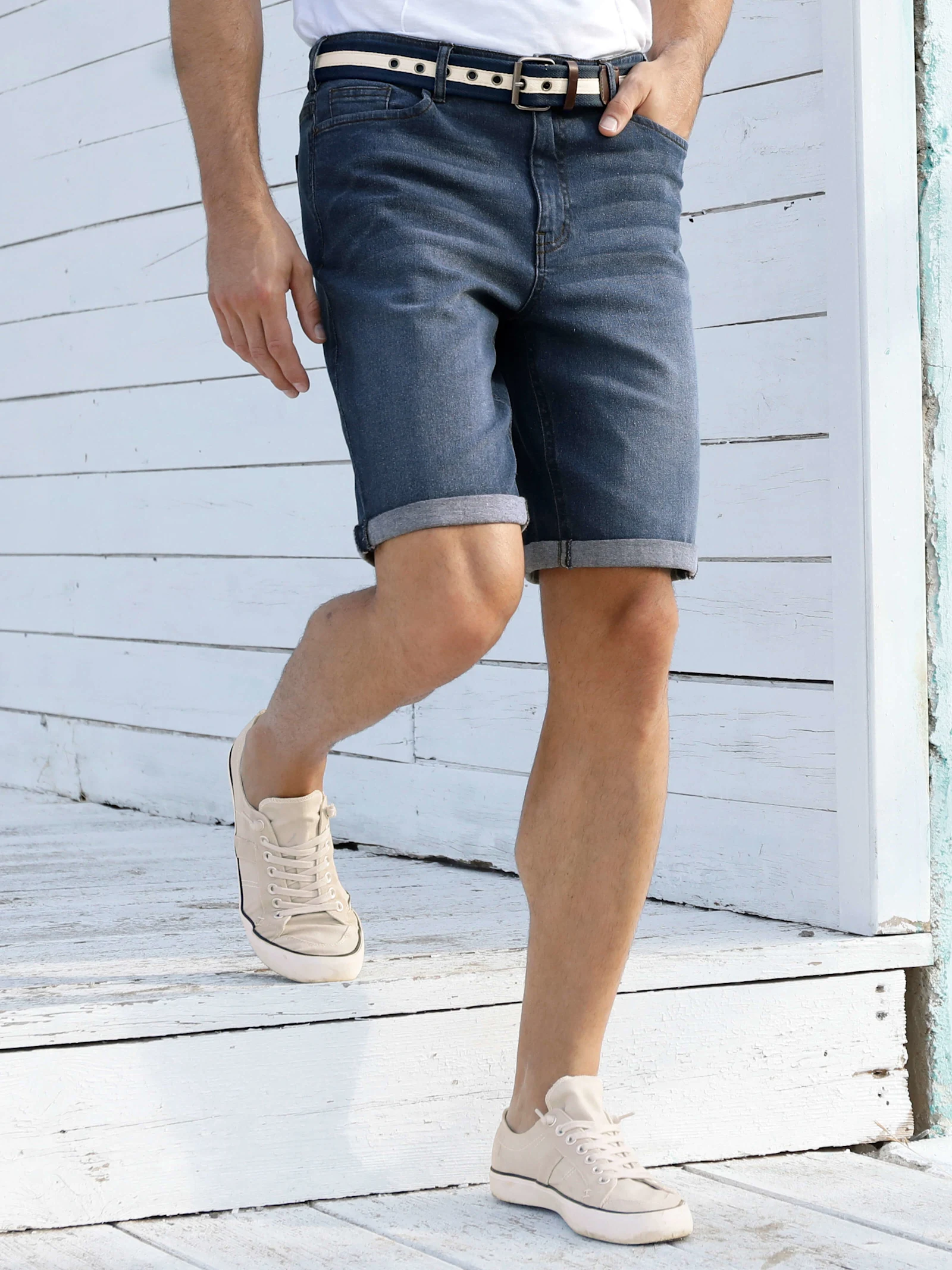 Ein männliches Model trägt dunkelblaue Jeansshorts mit Gürtel, helles Shirt und beigefarbene Sneaker, läuft auf einer weißen Holzterrasse.