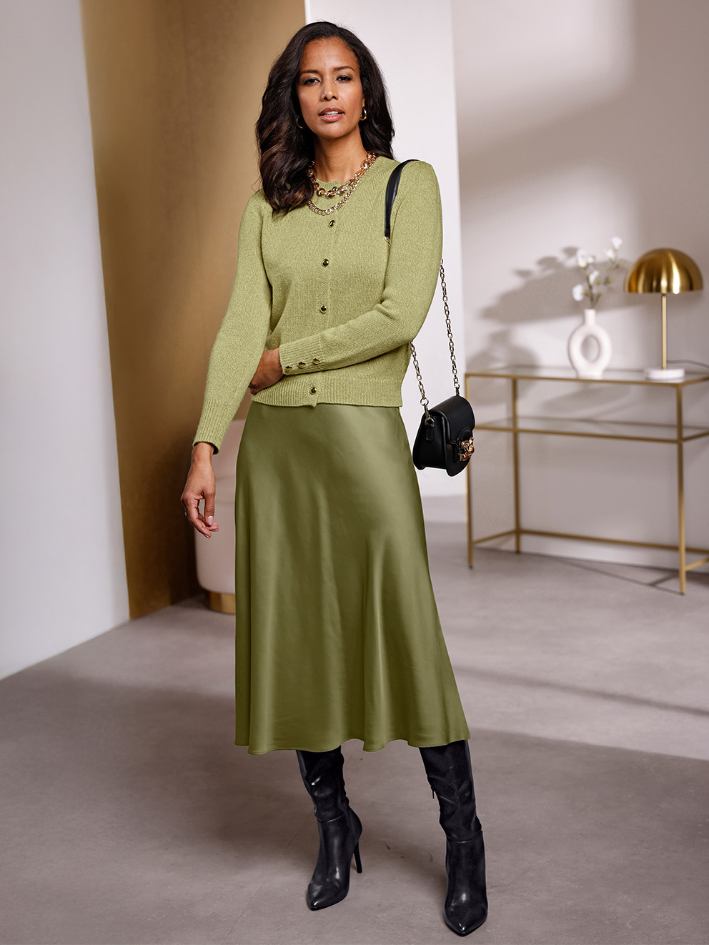Ein weibliches Model trägt eine schilf-melierte Strickjacke mit Schmuckknöpfen und einen olivgrünen Satinrock in ausgestellter Form, kombiniert mit schwarzen Stiefeln und einer schwarzen Tasche, in einem modernen, hellen Raum.
