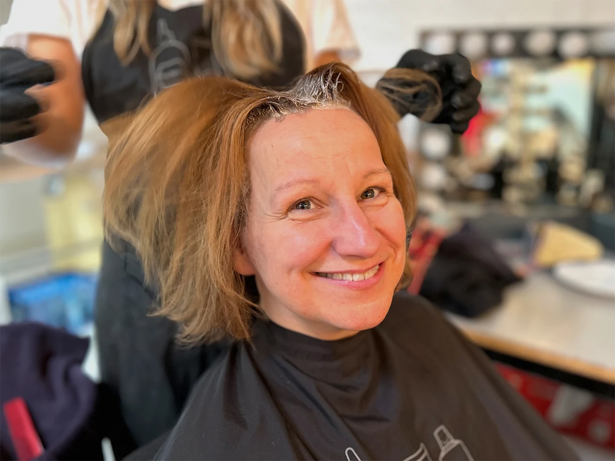 Eine Frau sitzt mit einem schwarzen Friseurumhang, während eine Person mit schwarzen Handschuhen die Haare blondiert. Eine Frau sitzt mit einem schwarzen Friseurumhang, während eine Person mit schwarzen Handschuhen die Haare blondiert.