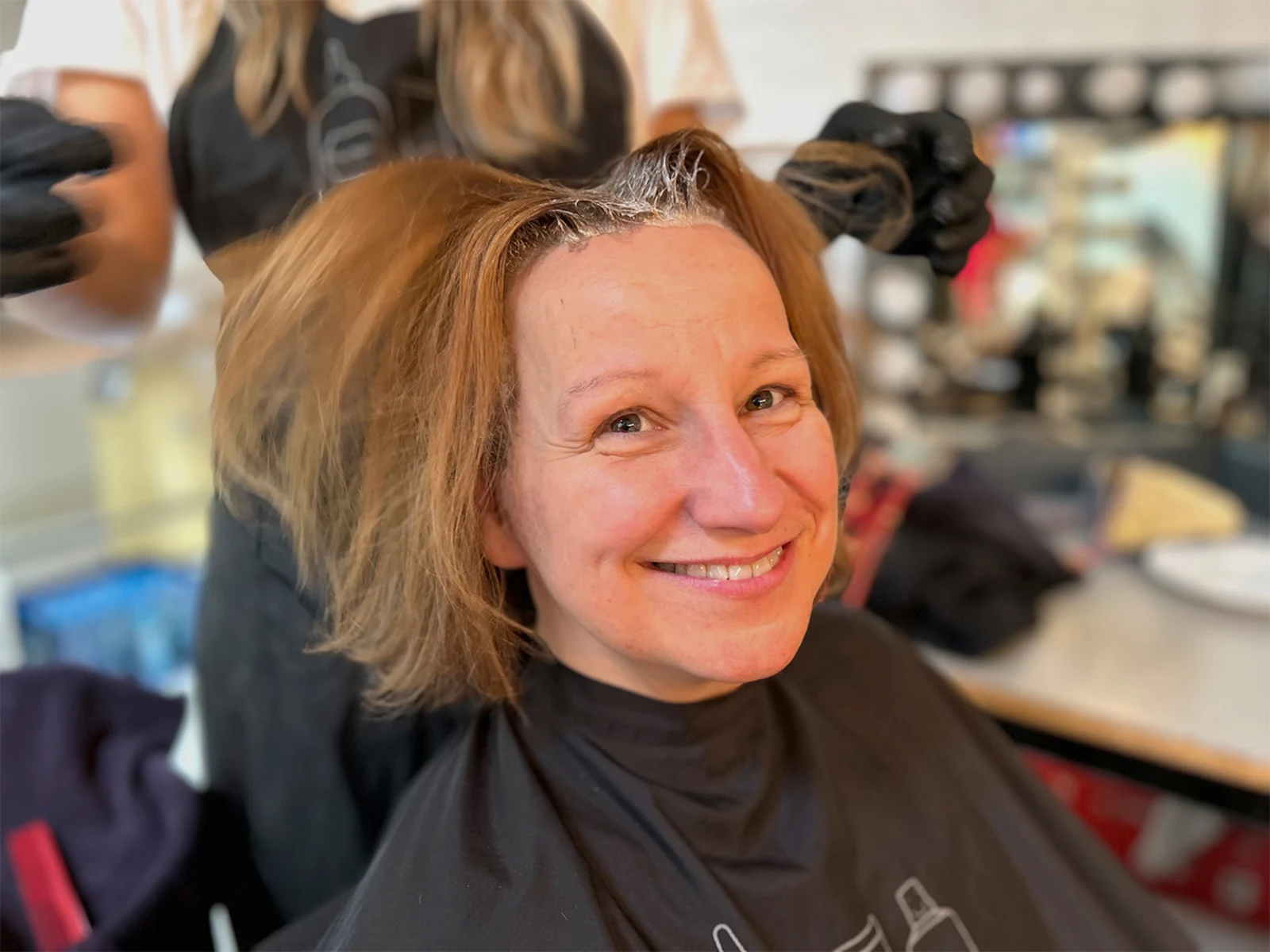 Eine Frau sitzt mit einem schwarzen Friseurumhang, während eine Person mit schwarzen Handschuhen die Haare blondiert. Eine Frau sitzt mit einem schwarzen Friseurumhang, während eine Person mit schwarzen Handschuhen die Haare blondiert.