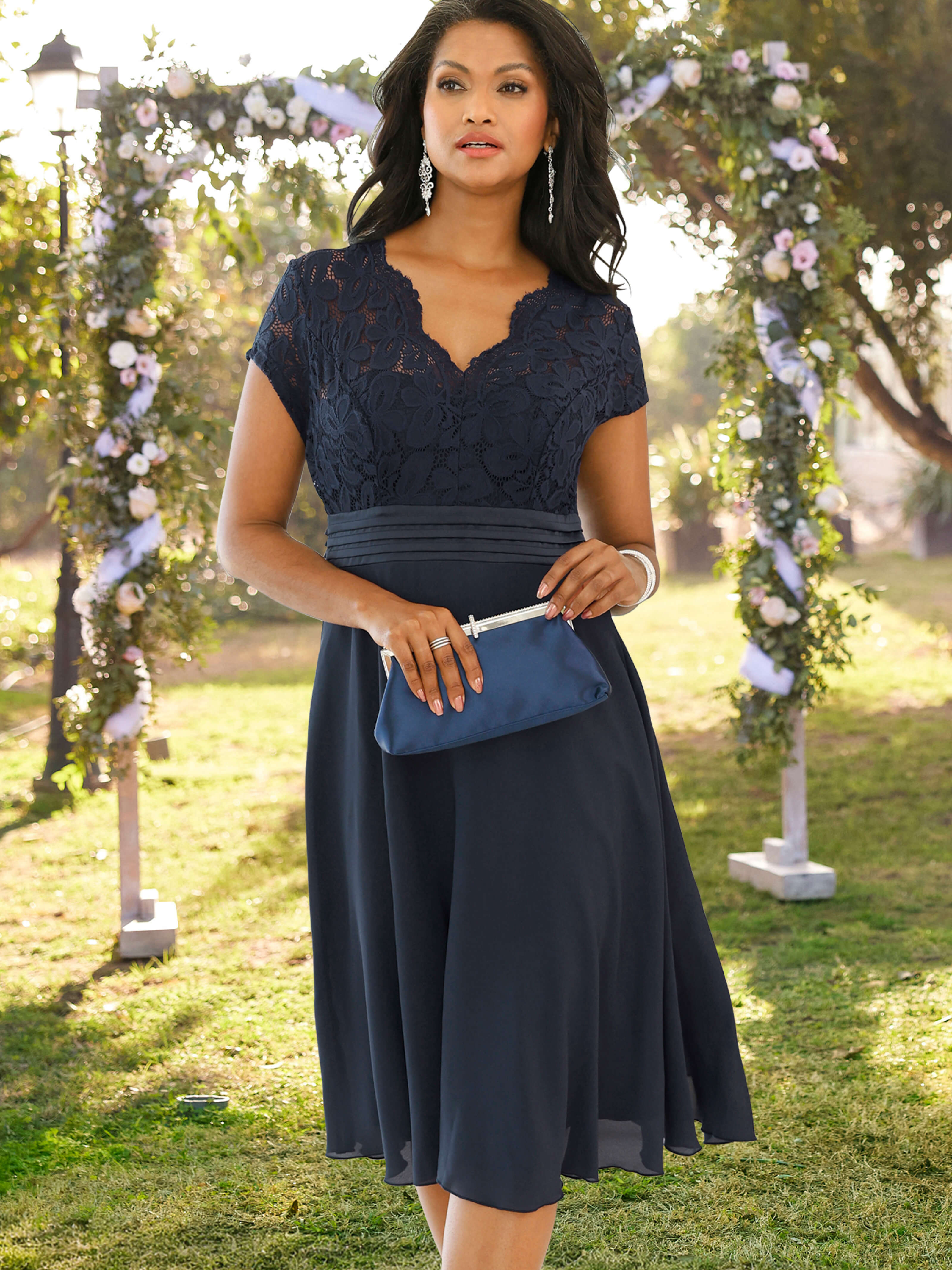 Das Bild zeigt ein Model in einem marinefarbenen Cocktailkleid mit kunstvollem Spitzeneinsatz, die eine blaue Clutch in der Hand hält und im Freien auf einer Wiese, vor einem mit Blumen verzierten Bogen steht.