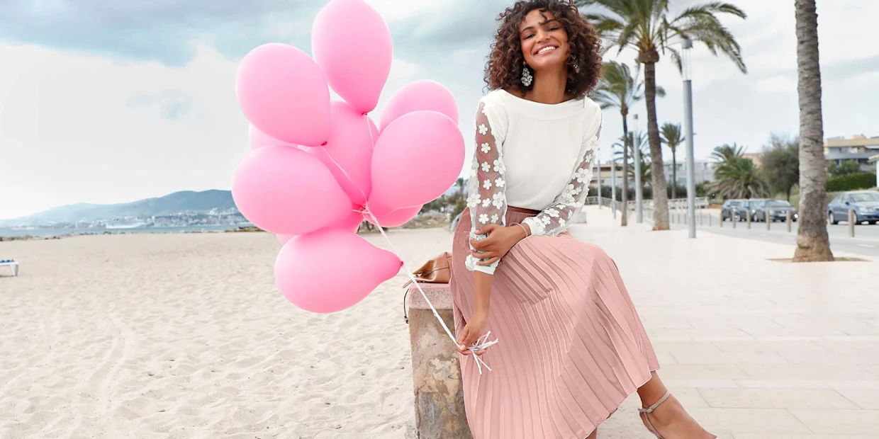 Frau in elegantem Outfit mit weißer Bluse und rosa Plisseerock, sitzt auf einer Steinbank am Strand, hält pinke Luftballons, im Hintergrund Palmen und Meer. Frau in elegantem Outfit mit weißer Bluse und rosa Plisseerock, sitzt auf einer Steinbank am Strand, hält pinke Luftballons, im Hintergrund Palmen und Meer.