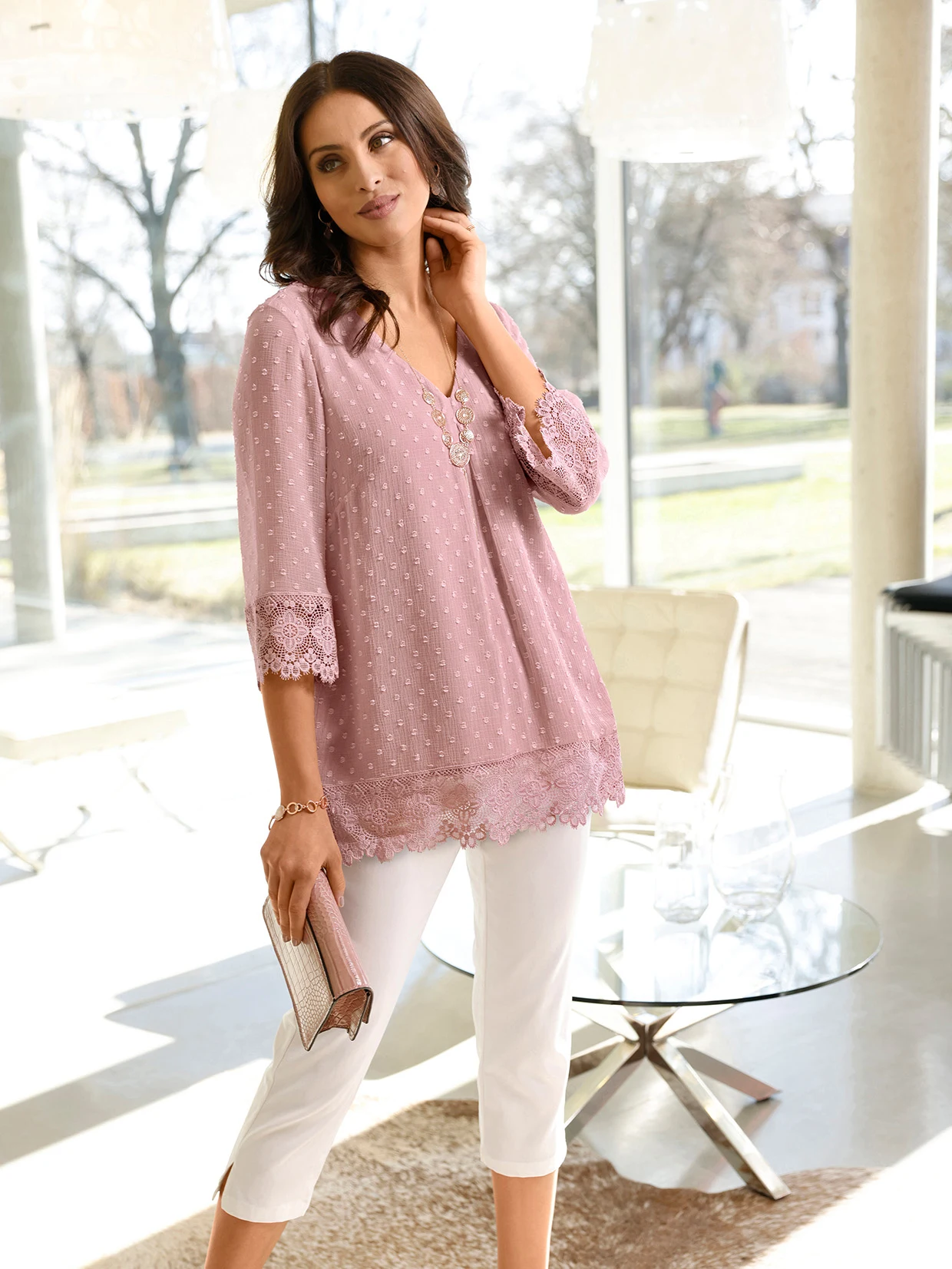Vrouw draagt een lichtroze tuniek met kanten details en een witte broek, terwijl ze een clutch vasthoudt in een moderne, lichte kamer.