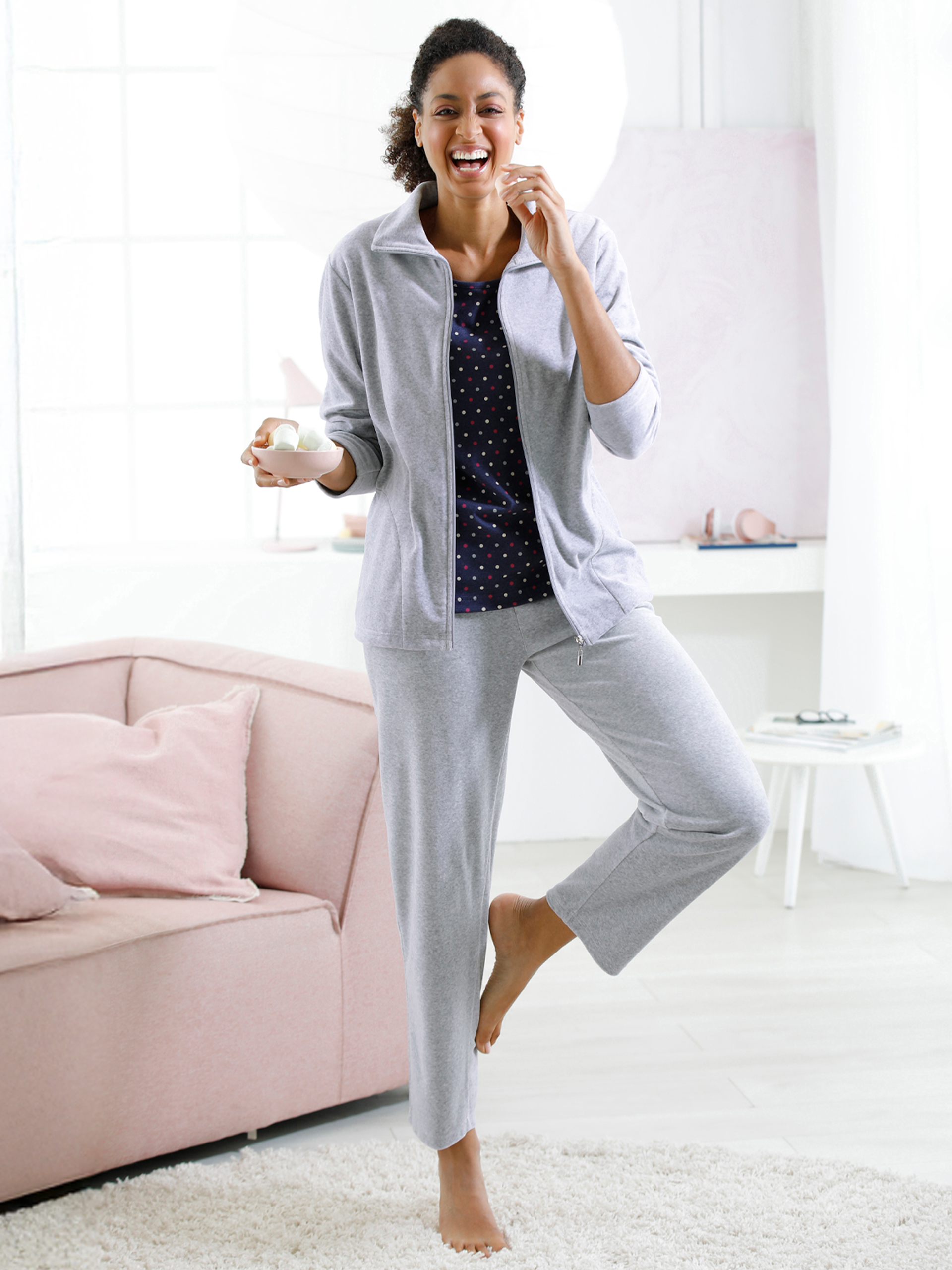 Ein Model steht barfuß in einem hellen Raum, trägt graue Freizeitkleidung und hält eine Tasse in der Hand.