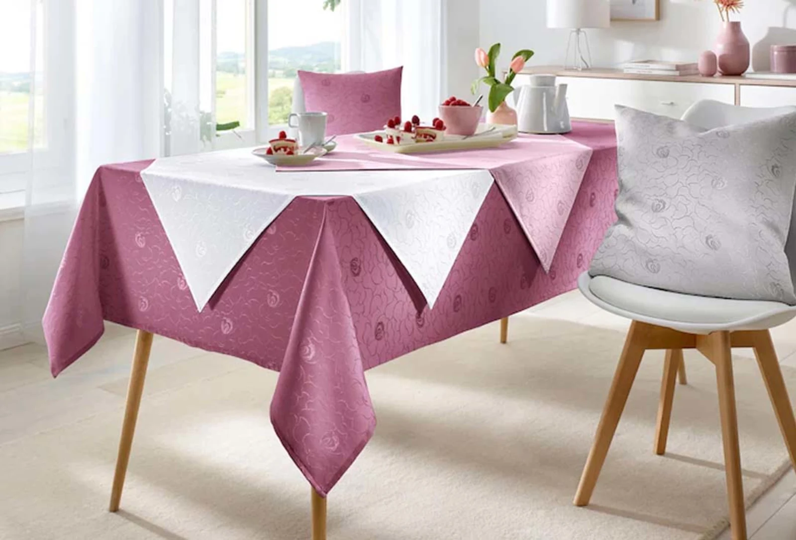 Eettafel met roze tafelkleed en wit tafelloper, gedecoreerd met borden, kopjes en een schaal met fruit, naast een stoel met bijpassend decoratief kussen. Eettafel met roze tafelkleed en wit tafelloper, gedecoreerd met borden, kopjes en een schaal met fruit, naast een stoel met bijpassend decoratief kussen.