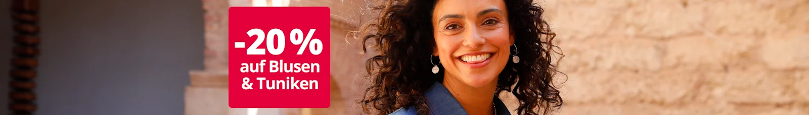Das Bild zeigt eine Frau mit lockigen Haaren in einem blauen Kleid, die einen bunten Blumenstrauß vor einer mediterranen Steinmauer hält. Auf roter Fläche steht der Text '-20 % auf Blusen & Tuniken'.