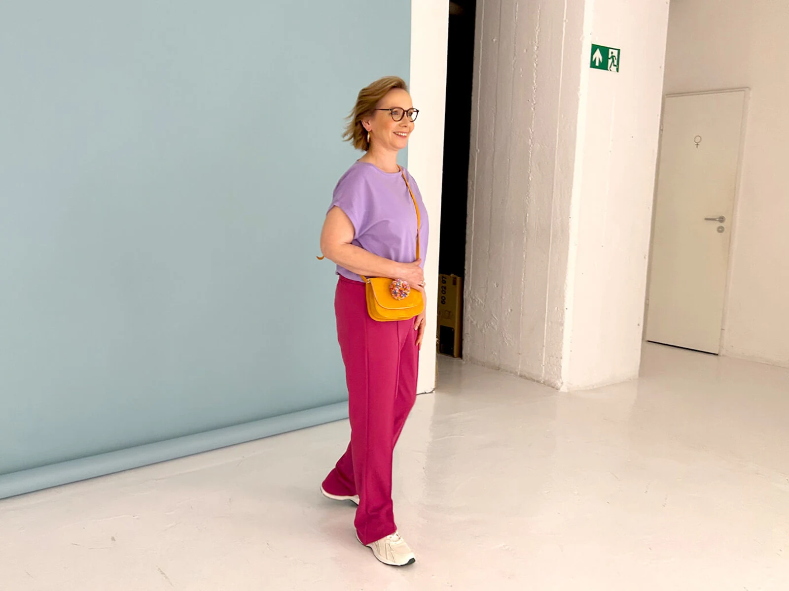 Eine Frau in lila Oberteil, roten Hosen und gelber Tasche, kombiniert mit weißen Sneakers, steht vor einer hellblauen Leinwand in einem hellen Raum. Eine Frau in lila Oberteil, roten Hosen und gelber Tasche, kombiniert mit weißen Sneakers, steht vor einer hellblauen Leinwand in einem hellen Raum.