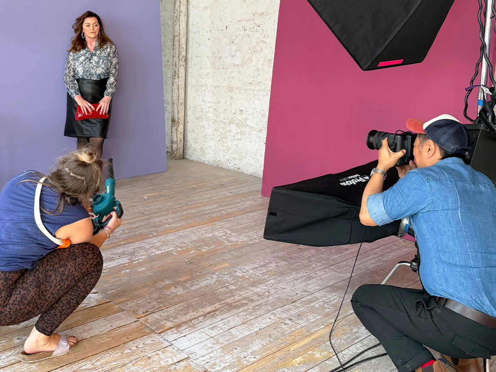 Eine Frau steht vor lilafarbener Wand, hält eine rote Clutch. Zwei Personen arbeiten: Eine mit Kamera, eine mit Windmaschine. Eine Frau steht vor lilafarbener Wand, hält eine rote Clutch. Zwei Personen arbeiten: Eine mit Kamera, eine mit Windmaschine.