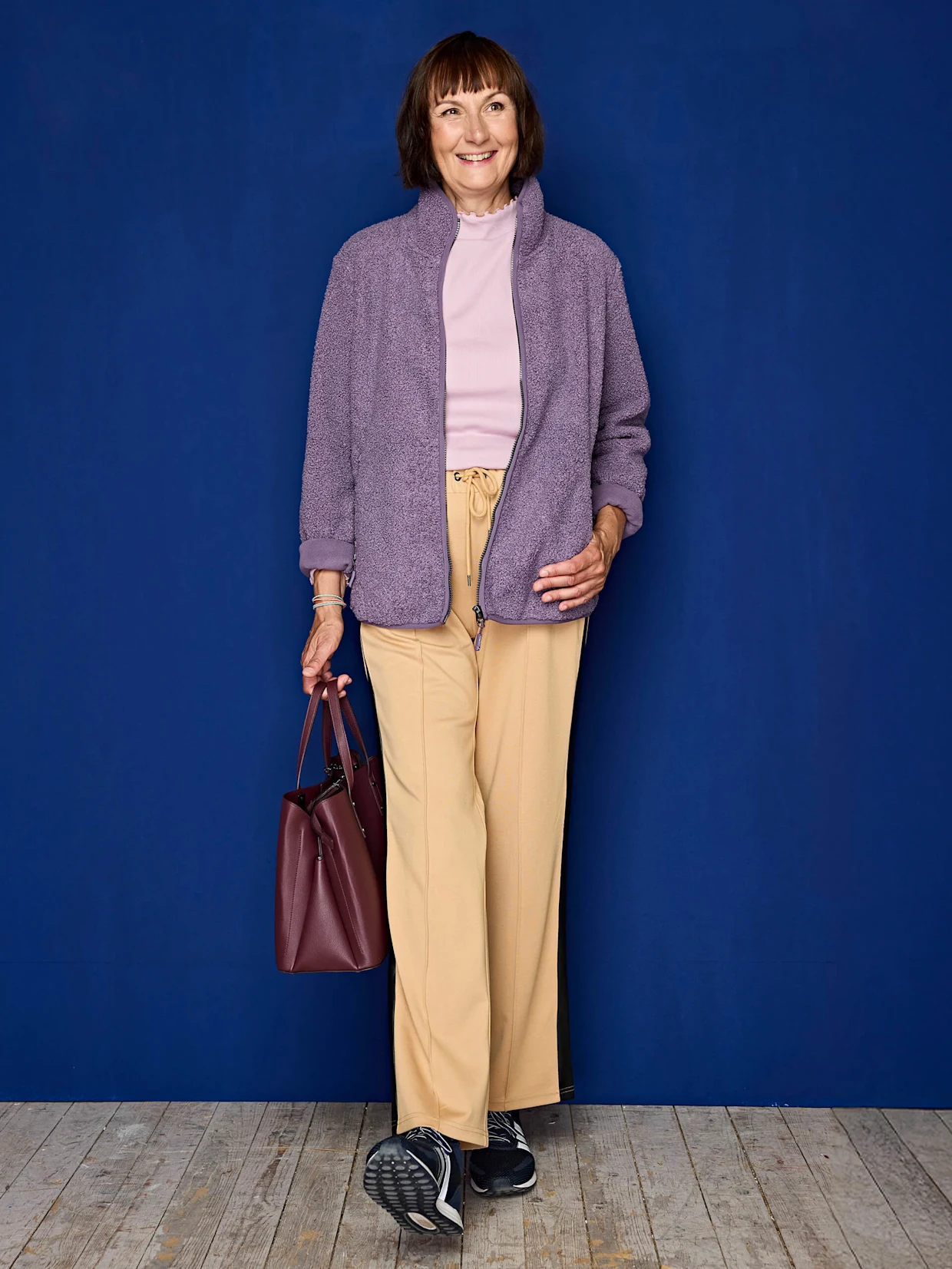 Eine Frau trägt eine fliederfarbene Fleecejacke, ein hellrosa Oberteil, beige Hosen mit Kordelzug, schwarze Sportschuhe und hält eine dunkelrote Tasche vor blauem Hintergrund.