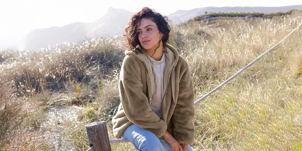Ein Model trägt eine beige, flauschige Kapuzenjacke über einem hellen Hoodie und blaue Jeans. Sie sitzt draußen auf einem Seil mit Gras und Bergen im Hintergrund.