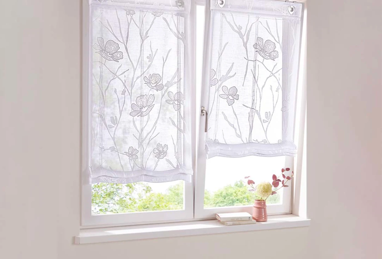 Zwei weiße Fenster mit halbtransparenten Rollos, die mit floralen Mustern verziert sind; auf der Fensterbank stehen eine rosa Vase mit Dekoblumen und zwei Bücher. Zwei weiße Fenster mit halbtransparenten Rollos, die mit floralen Mustern verziert sind; auf der Fensterbank stehen eine rosa Vase mit Dekoblumen und zwei Bücher.