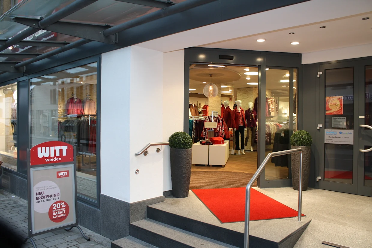 Eingangsbereich einer Witt Weiden Filiale mit rotem Teppich, Schaufenster mit Kleidung und Werbeschild zur Neueröffnung mit 20% Rabatt.
