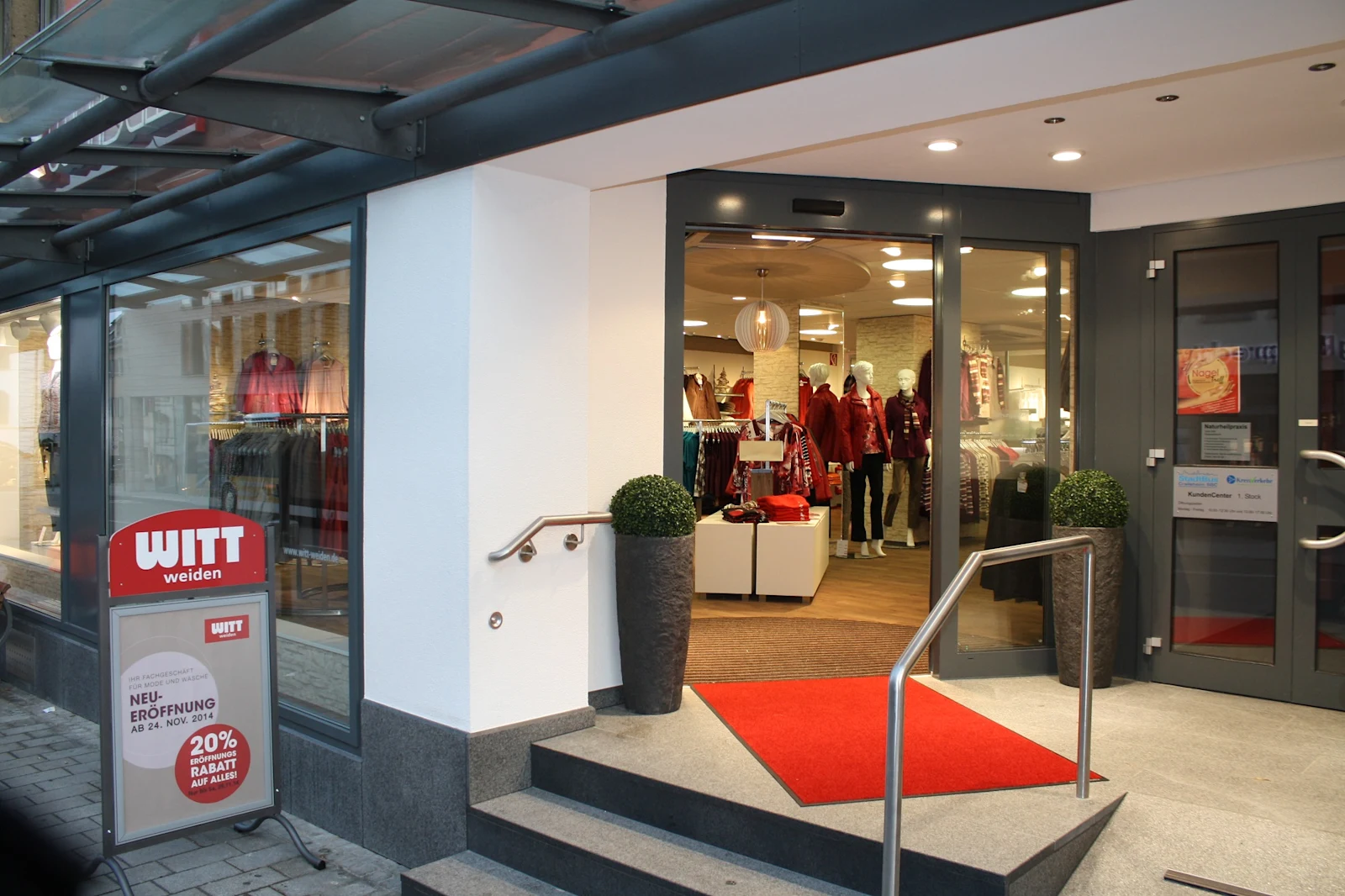 Eingangsbereich einer Witt Weiden Filiale mit rotem Teppich, Schaufenster mit Kleidung und Werbeschild zur Neueröffnung mit 20% Rabatt.