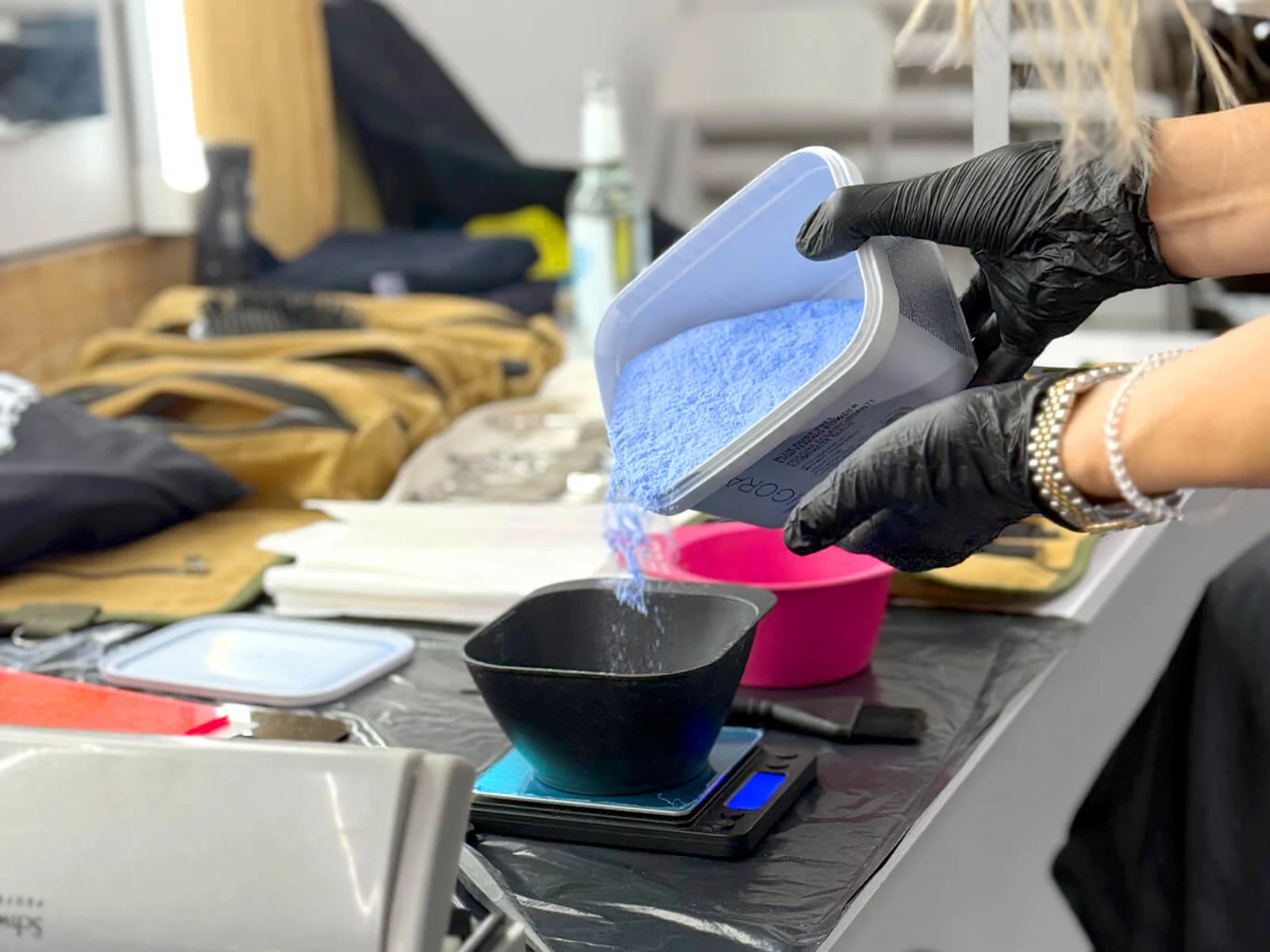 Eine Person mit schwarzen Handschuhen gießt blauen Puder in eine Schüssel auf einer Waage. Im Hintergrund sind verschiedene Utensilien zu sehen. Eine Person mit schwarzen Handschuhen gießt blauen Puder in eine Schüssel auf einer Waage. Im Hintergrund sind verschiedene Utensilien zu sehen.