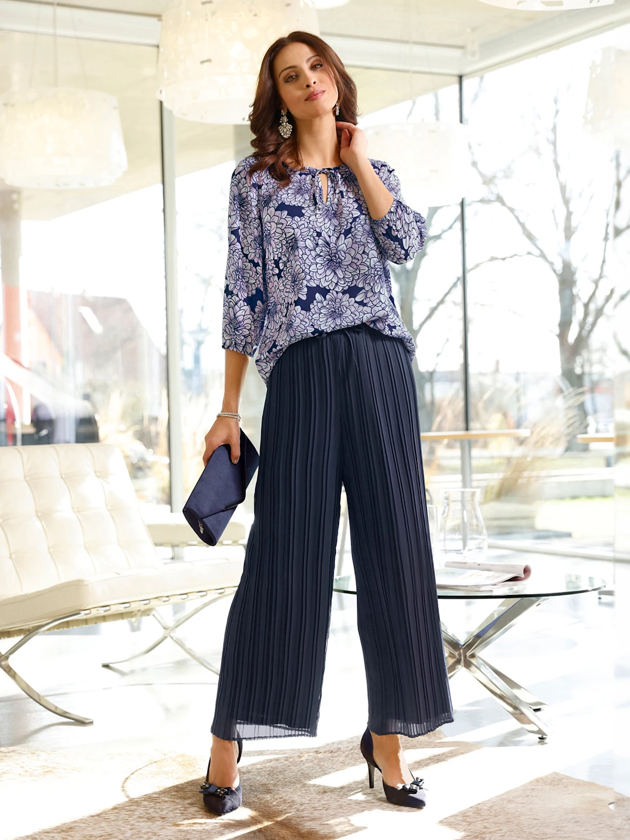 Frau trägt eine dunkelblaue, gemusterte Bluse mit floralen Details, kombiniert mit weiter, plissierter Hose und passenden High-Heels, dazu eine dunkelblaue Clutch in einer modernen Wohnumgebung.