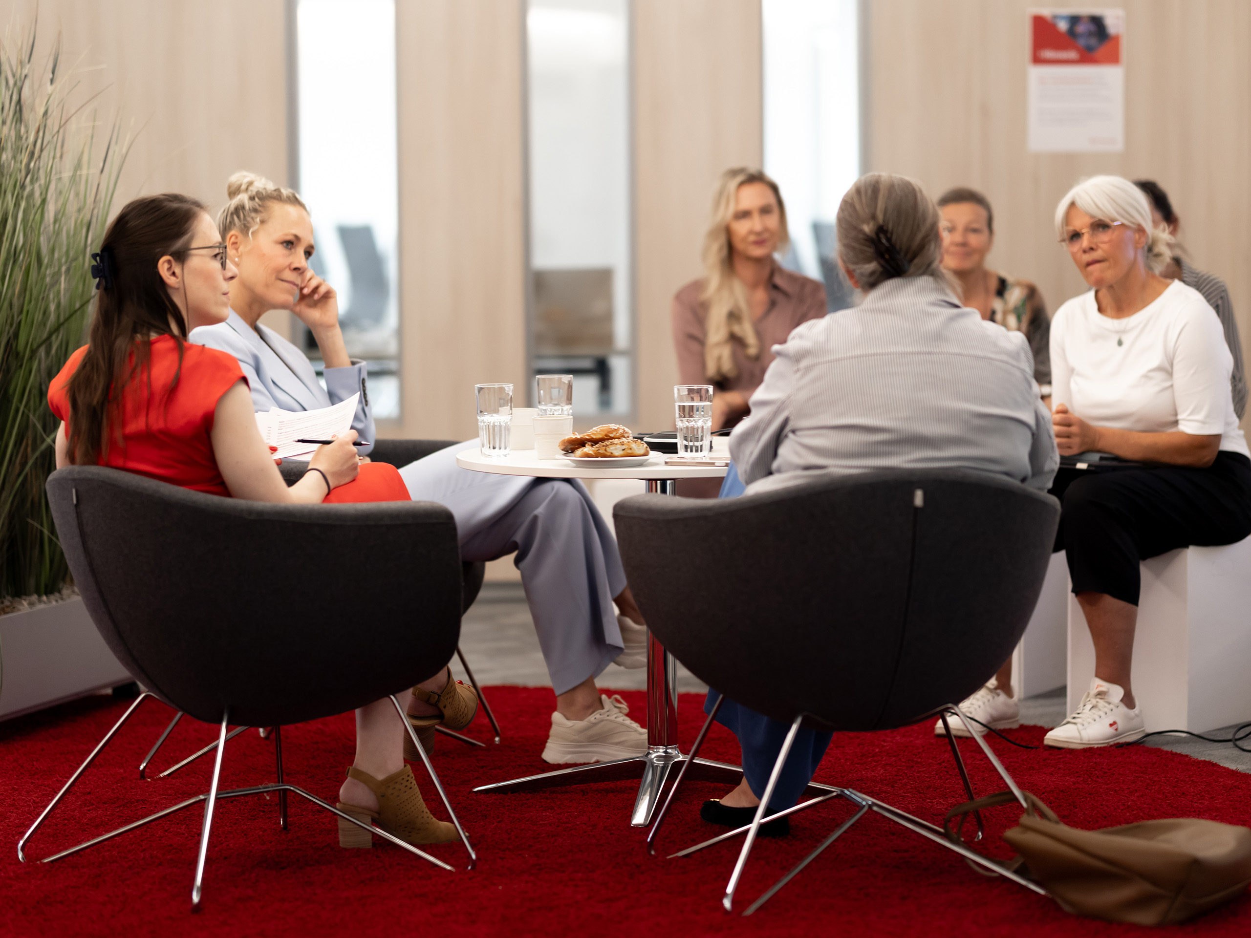 Gruppe von Frauen in Stühlen um einen kleinen Tisch mit Gläsern und Gebäck in einem Besprechungsraum.