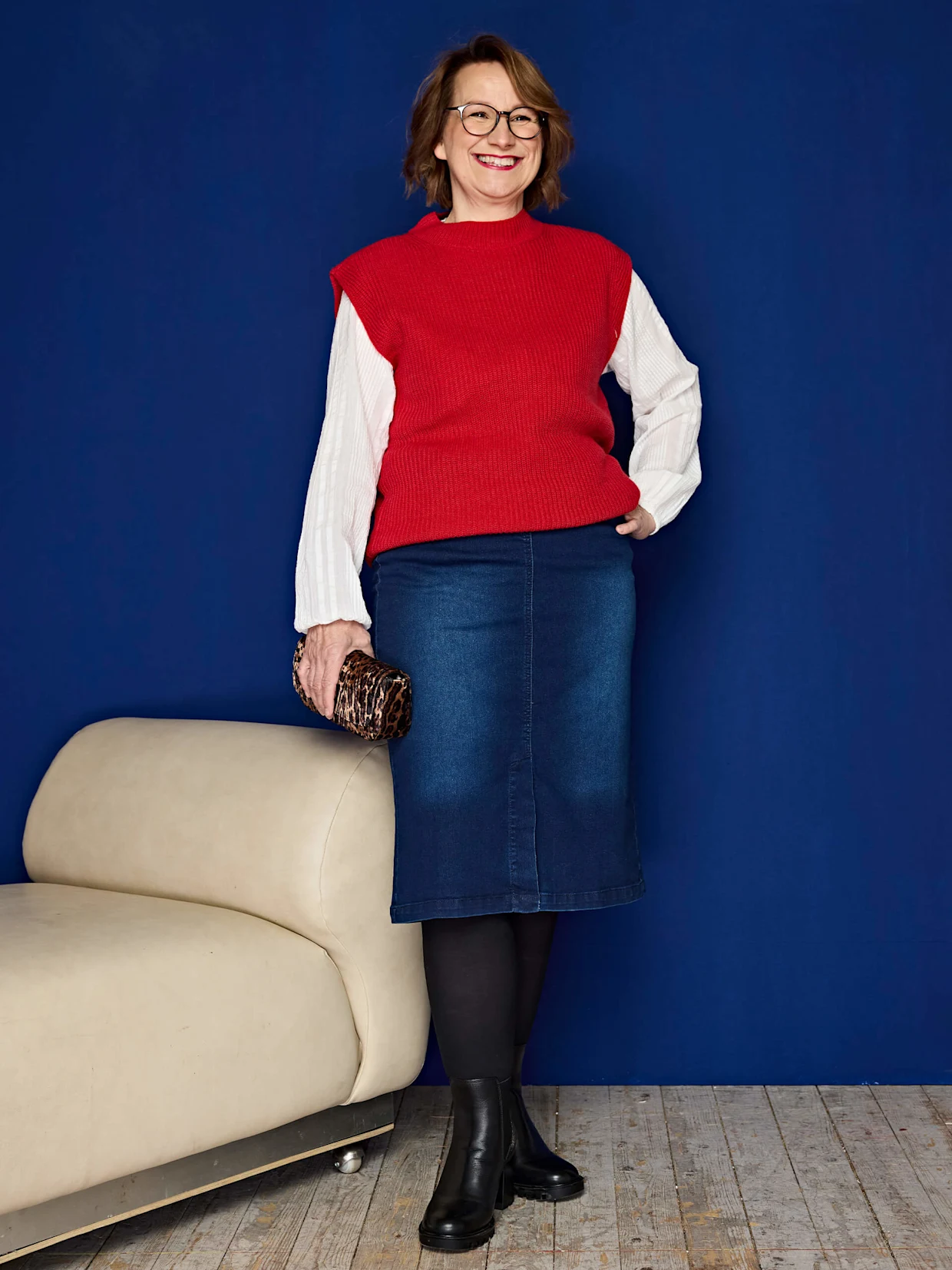 Eine Frau trägt einen roten Strickpullunder über einem weißen Langarmshirt, blauen Jeansrock, schwarze Strumpfhose und schwarze Stiefeletten, hält eine kleine Tasche. Der Hintergrund ist blau.