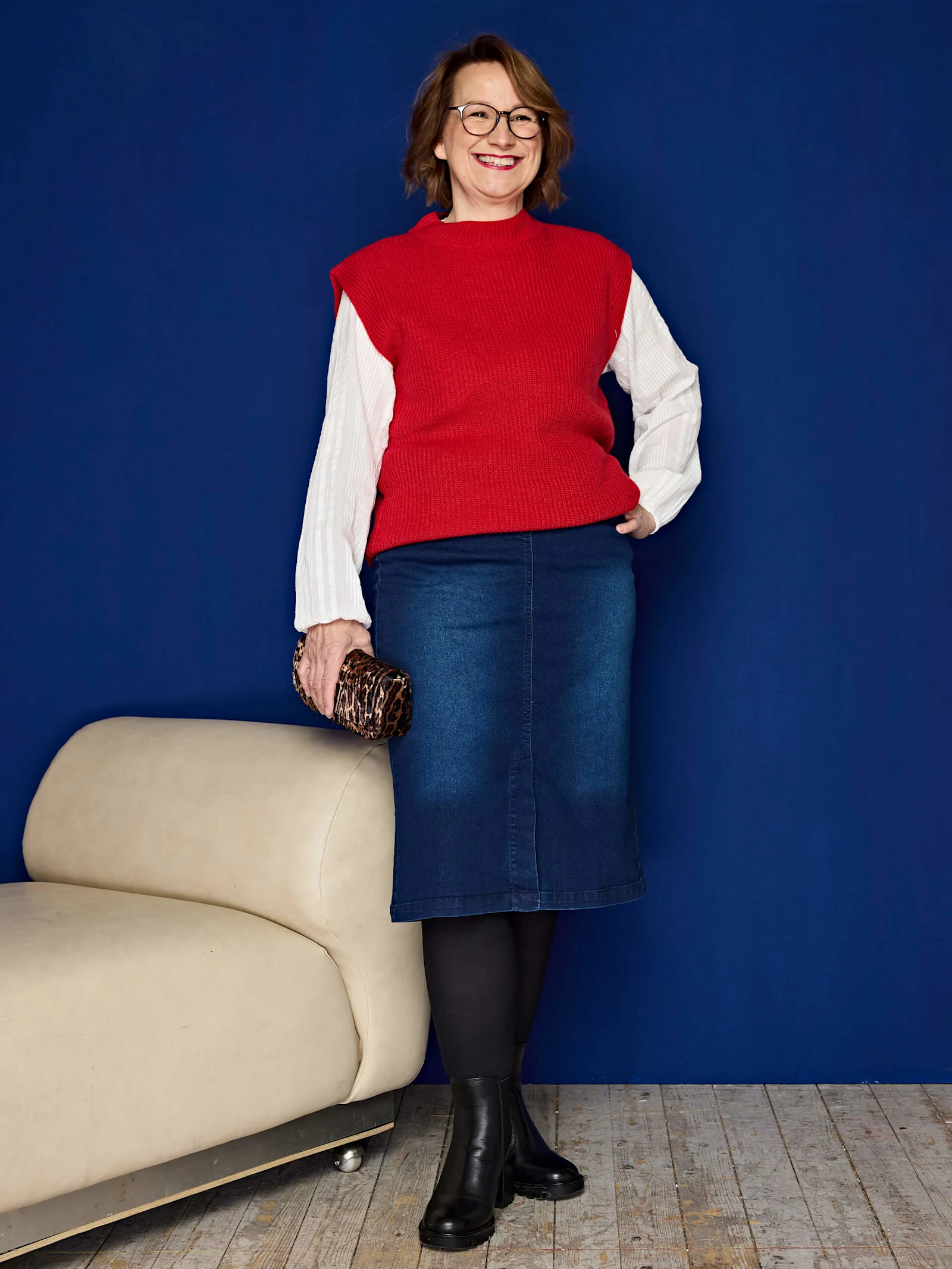 Eine Frau trägt einen roten Strickpullunder über einem weißen Langarmshirt, blauen Jeansrock, schwarze Strumpfhose und schwarze Stiefeletten, hält eine kleine Tasche. Der Hintergrund ist blau.