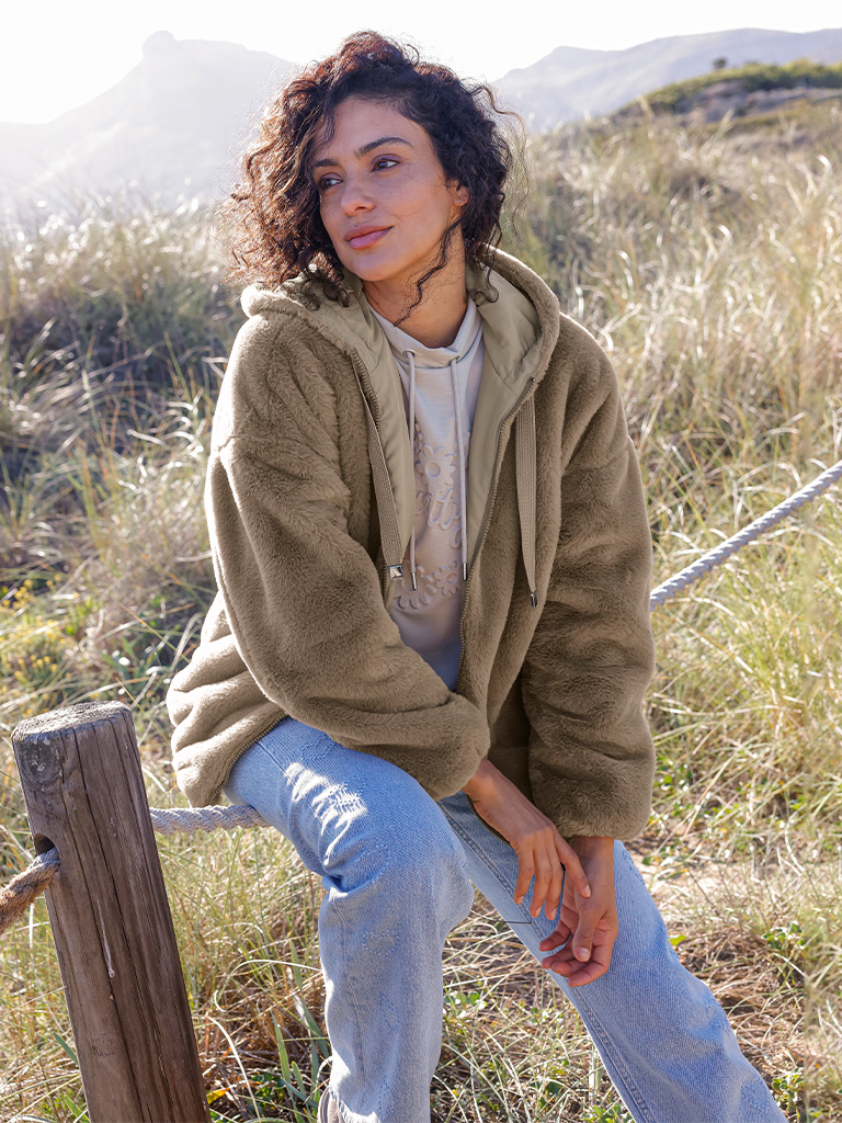 Ein Model trägt eine schlammfarbene Teddyfelljacke mit Kapuze und helle Jeans, sitzt draußen auf einem Holzzaun.