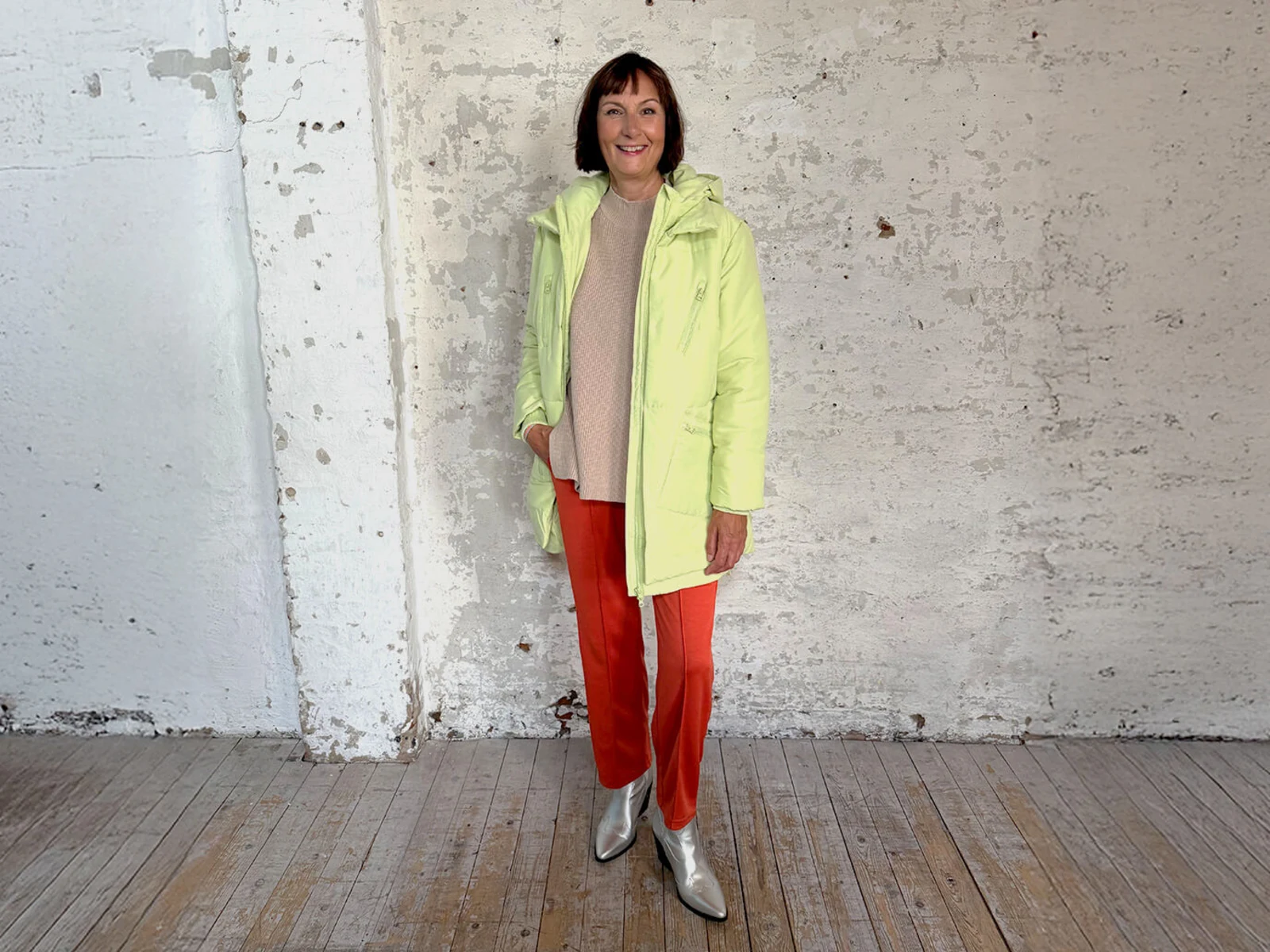 Eine Frau trägt eine helle, wattierte Jacke, einen beigen Pullover, orangefarbene Hosen und silberne Stiefeletten vor einer weißen, rauen Wand. Eine Frau trägt eine helle, wattierte Jacke, einen beigen Pullover, orangefarbene Hosen und silberne Stiefeletten vor einer weißen, rauen Wand.