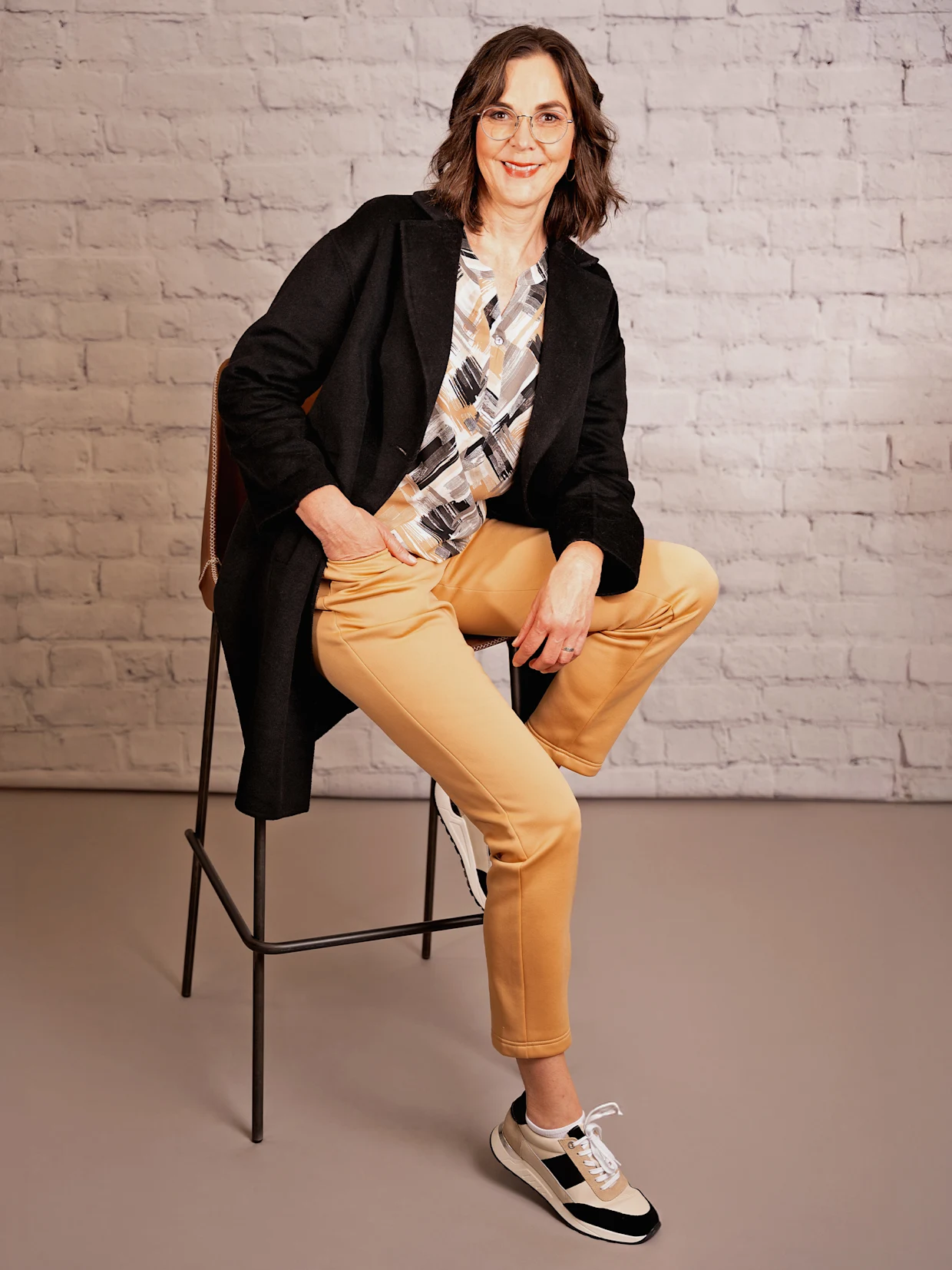Eine braunhaarige Frau sitzt auf einem Hocker und trägt eine ecru-schwarz-bedruckte Schlupfbluse mit elastischem Saum, eine camelfarbene Hose, einen schwarzen Kurzmantel in Doubleface-Qualität und beige-schwarze Sneaker. Der Hintergrund ist eine weiße Wand.