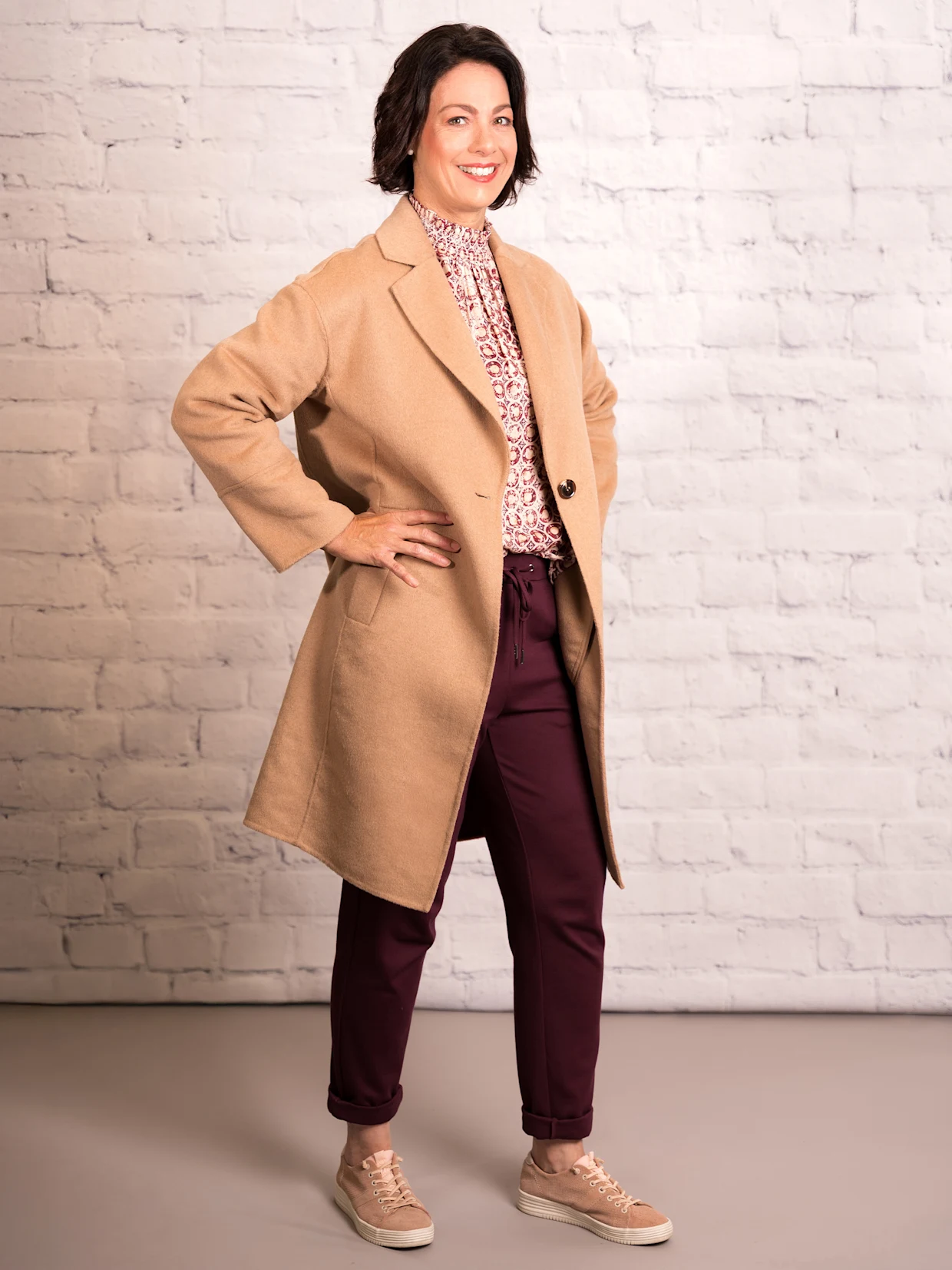 Eine dunkelhaarige Frau trägt einen camelfarbenen Kurzmantel in Doubleface-Qualität, eine elfenbein-mauve-bedruckte Bluse mit Nackenverschluss und burgundfarbene Jogg Pants in Interlock-Qualität. Der Hintergrund ist eine weiße Wand.