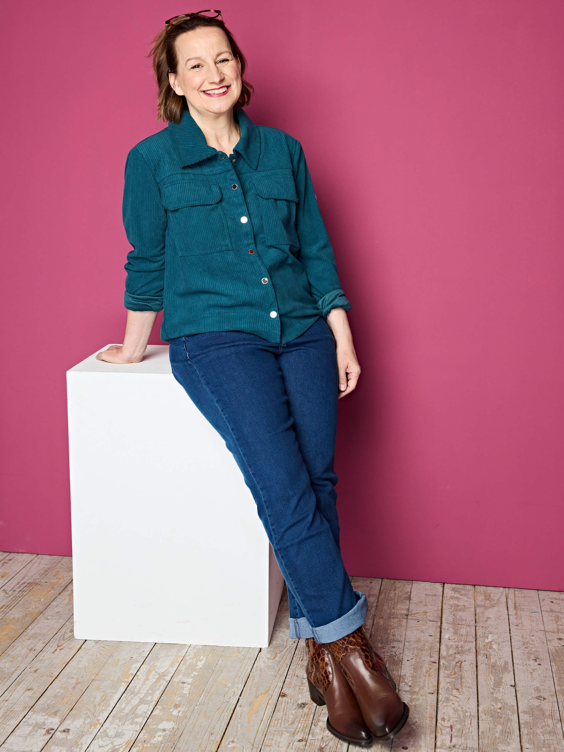 Eine Frau trägt eine petrolfarbene Cordjacke mit Knöpfen, blaue Jeans mit aufgekrempeltem Saum und braune Stiefeletten mit Muster vor pinkem Hintergrund.
