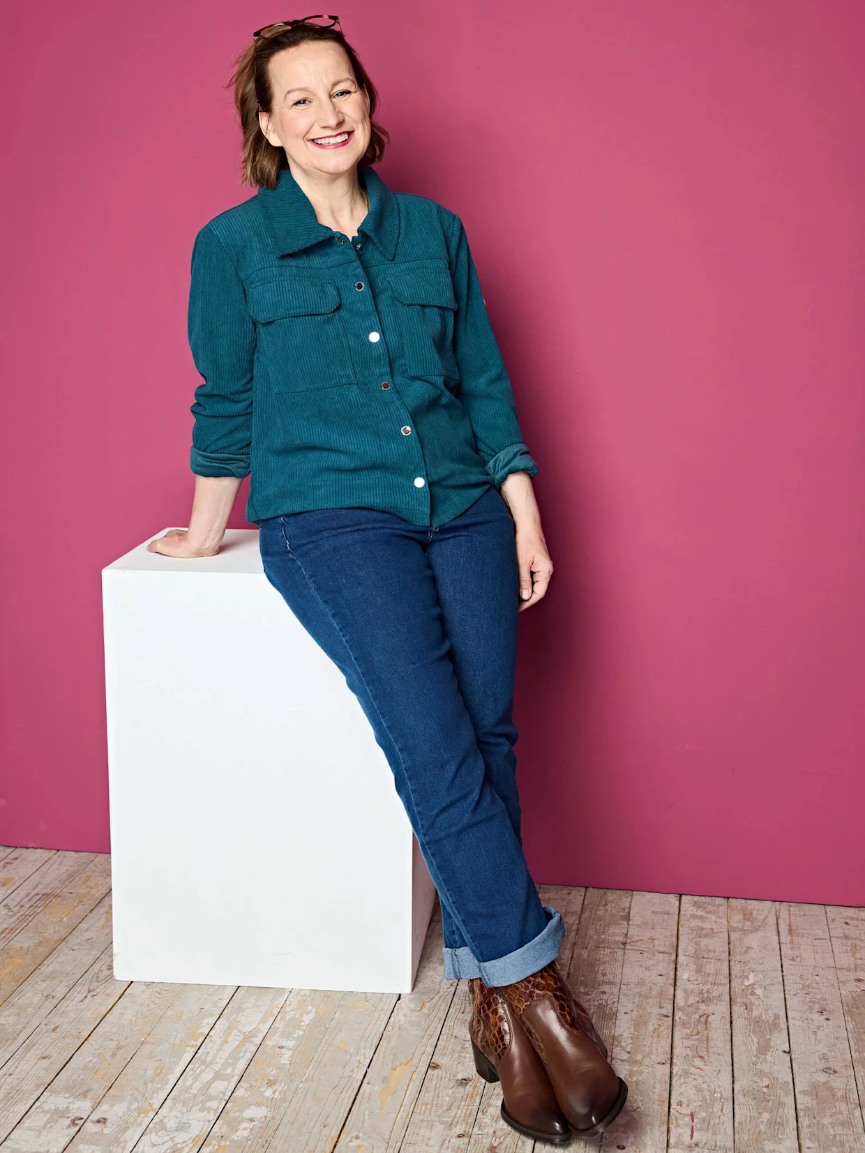 Eine Frau trägt eine petrolfarbene Cordjacke mit Knöpfen, blaue Jeans mit aufgekrempeltem Saum und braune Stiefeletten mit Muster vor pinkem Hintergrund.