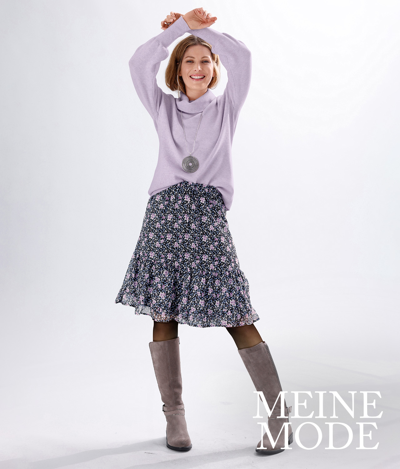 Frau trägt einen fliederfarbenen Pullover, einen gemusterten Rock mit Blumenprint und graue Stiefel.