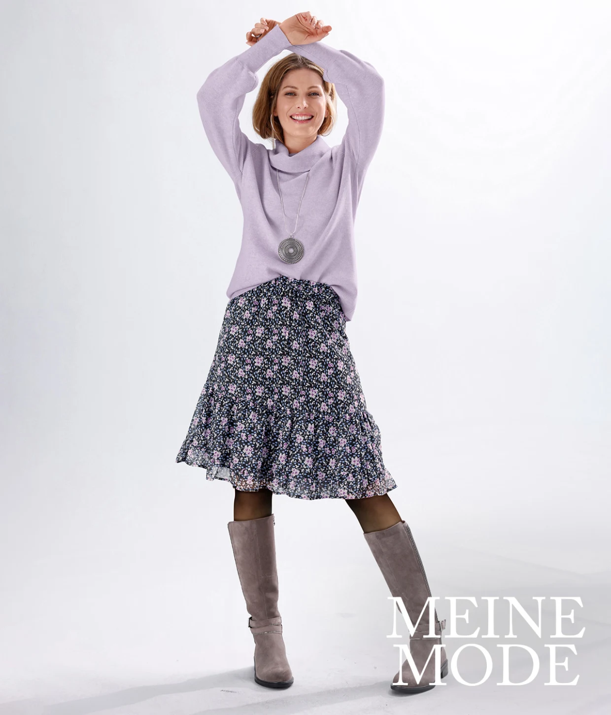 Frau trägt einen fliederfarbenen Pullover, einen gemusterten Rock mit Blumenprint und graue Stiefel.