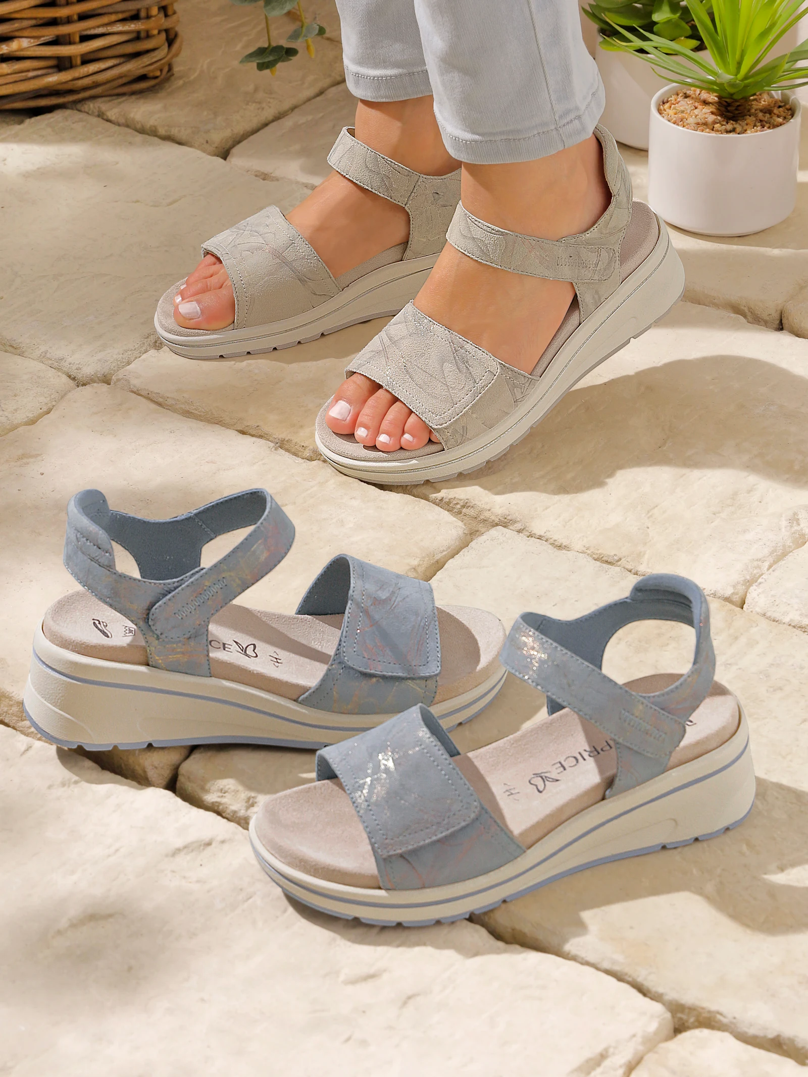 Ein Model trägt silberfarbene Plateausandalen mit Klettverschluss, daneben stehen ähnliche Sandalen in Hellblau, Pflanzen im Hintergrund.