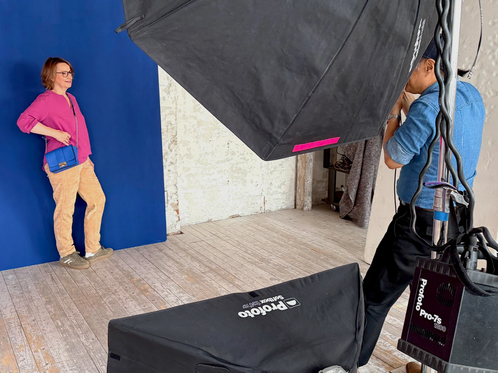 Eine Frau in pinker Bluse und beiger Hose posiert vor blauem Hintergrund, Fotograf mit Kamera und großer Softbox im Vordergrund. Eine Frau in pinker Bluse und beiger Hose posiert vor blauem Hintergrund, Fotograf mit Kamera und großer Softbox im Vordergrund.