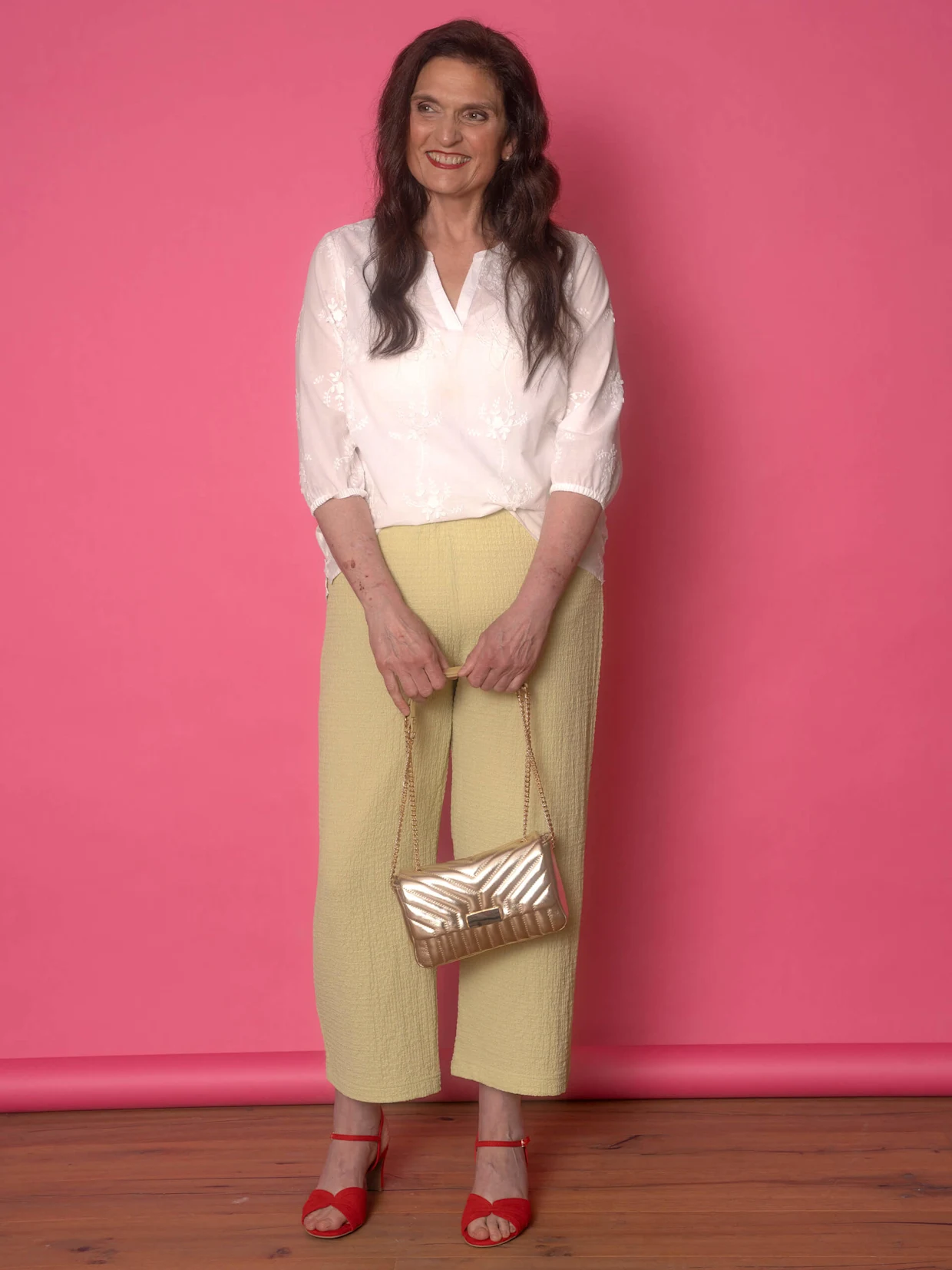Ein Model trägt eine weiße Bluse, gelbe Hose und rote Sandalen, während es eine goldene Handtasche hält, vor einem pinken Hintergrund.