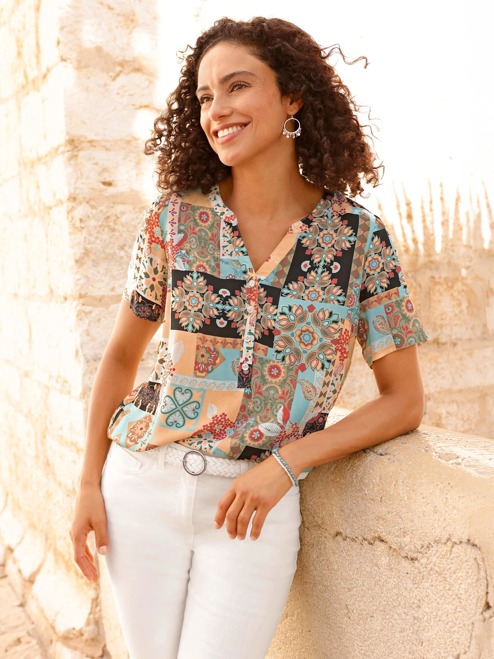 Ein Model lehnt an einer hellen Steinmauer, trägt eine bunte, gemusterte Bluse und weiße Hose.