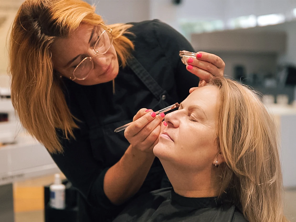 Eine blonde Frau sitzt und trägt einen schwarzen Friseurumhang während sie von einer anderen, rot-blonden Frau mit Brille geschminkt wird. Eine blonde Frau sitzt und trägt einen schwarzen Friseurumhang während sie von einer anderen, rot-blonden Frau mit Brille geschminkt wird.