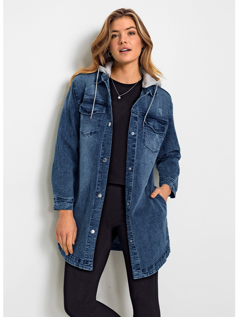 Ein Model trägt eine lange blaue Jeansjacke mit Kapuze, kombiniert mit schwarzer Kleidung, und steht vor einer weißen Wand.