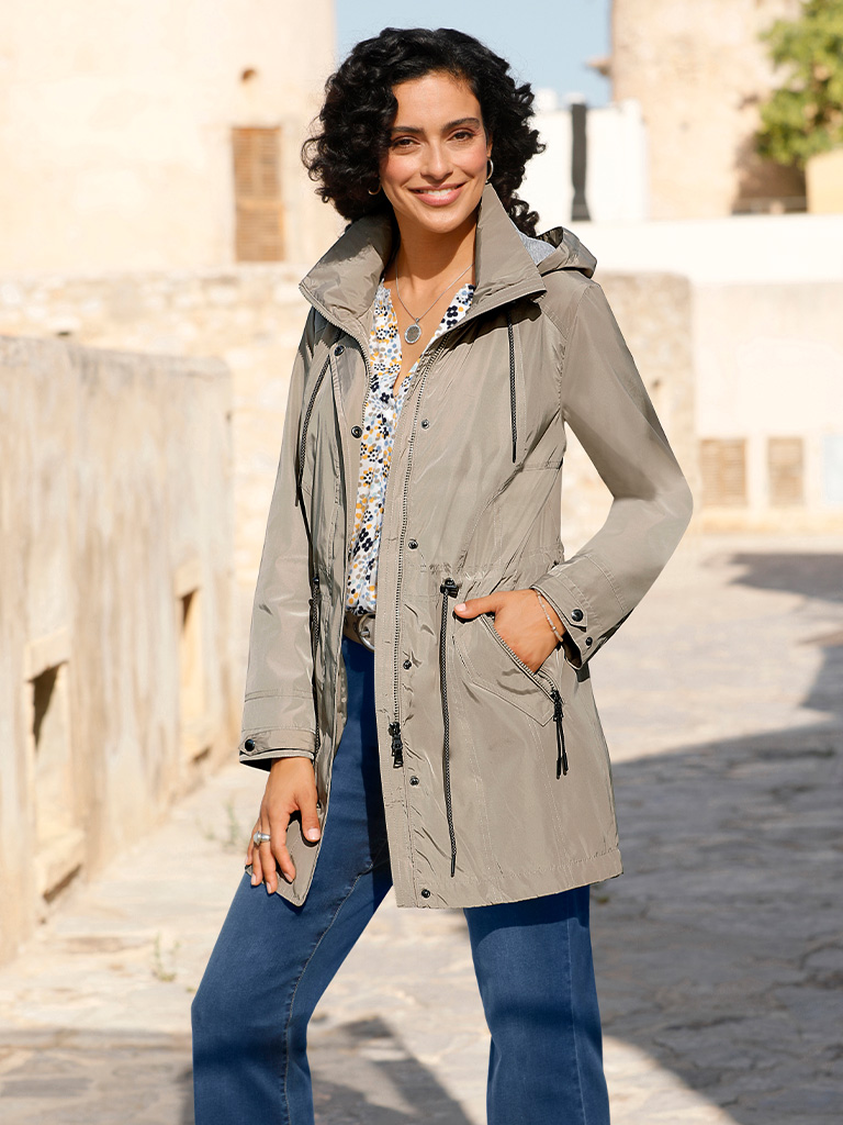 Ein Model trägt einen schlammfarbenen, knielangen Parka mit Kapuze, kombiniert mit blauen Jeans und einem gemusterten Oberteil, stehend im Freien.